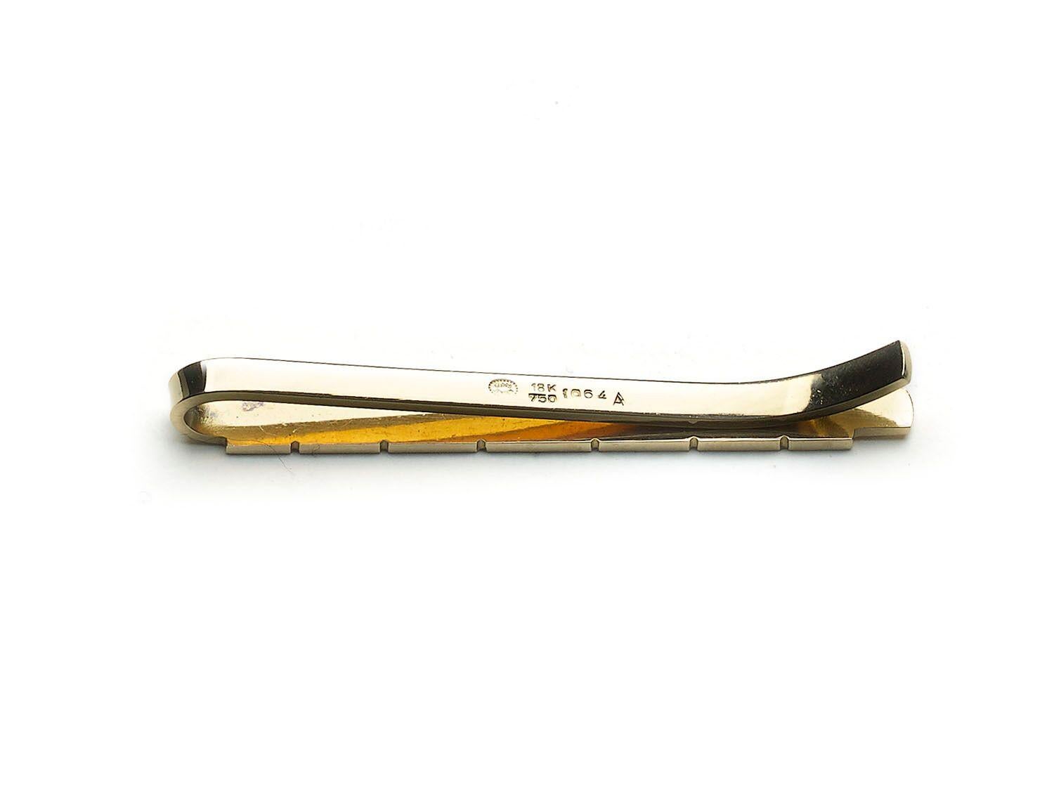 A Georg Jensen tie clip, with a row of graduating rectangles on a bar, designed by Henry Pilstrup, mounted in gold, with a post 1945 Georg Jensen maker's mark, in a dotted oval surround, stamped 18K 750, numbered 1064 A. Circa 1950.

Henry Pilstrup