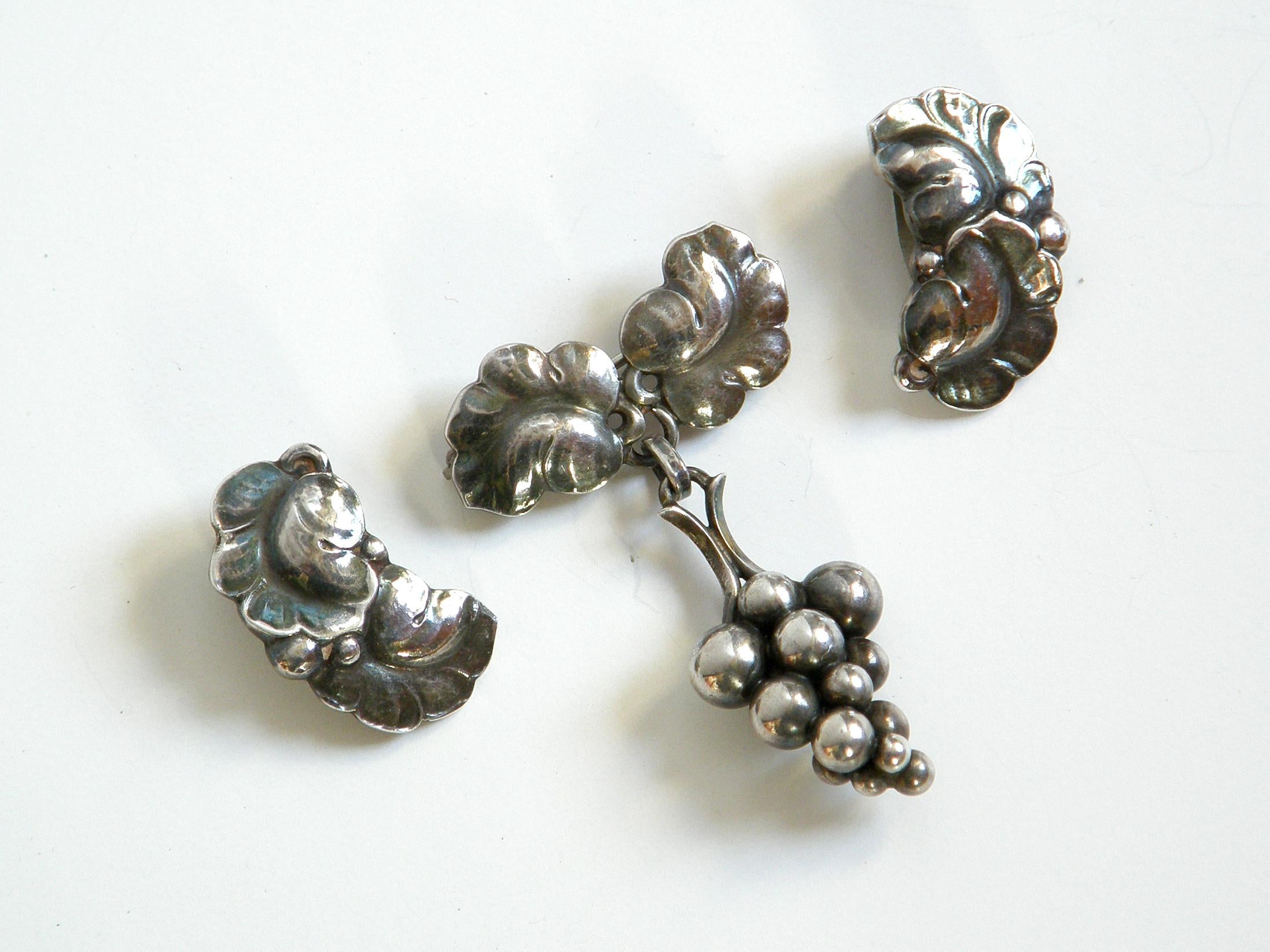 This sterling pin and earrings set is one of the classic designs of Georg Jensen. The design, known as Moonlight Grapes, features a brooch with a three-dimensional cluster of grapes hanging from curling leaves with a hammered finish. The matching