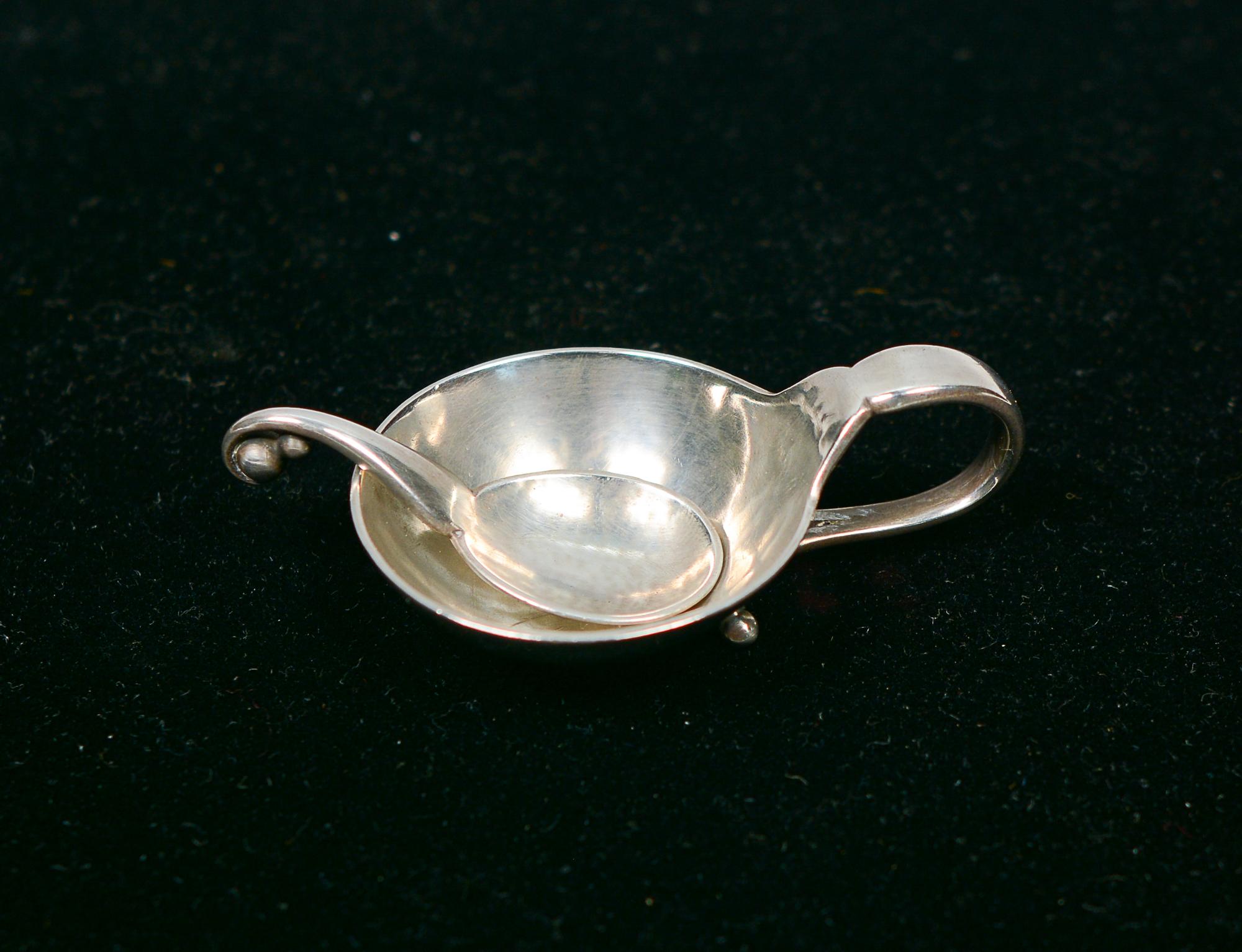 Sterling silver salt cellar and spoon model 110 by Georg Jensen. This is hand hammered. The handle folds around to the bottom and ends with two ball feet. The end of the spoon has a similar ball design. The mark indicates this was retailed in