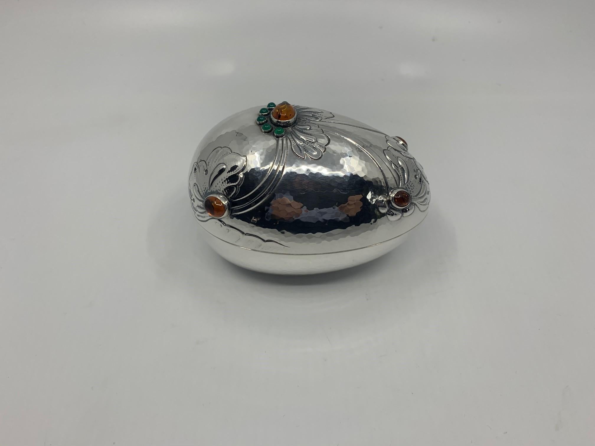 Oval Georg Jensen bonbonnière decorated with green agate and amber stones and beautiful hand-chased floral details. A small piece of hollowware that resembles a piece of jewelry.

Item number: 3522201

Stones: Green agate, Amber
Material: