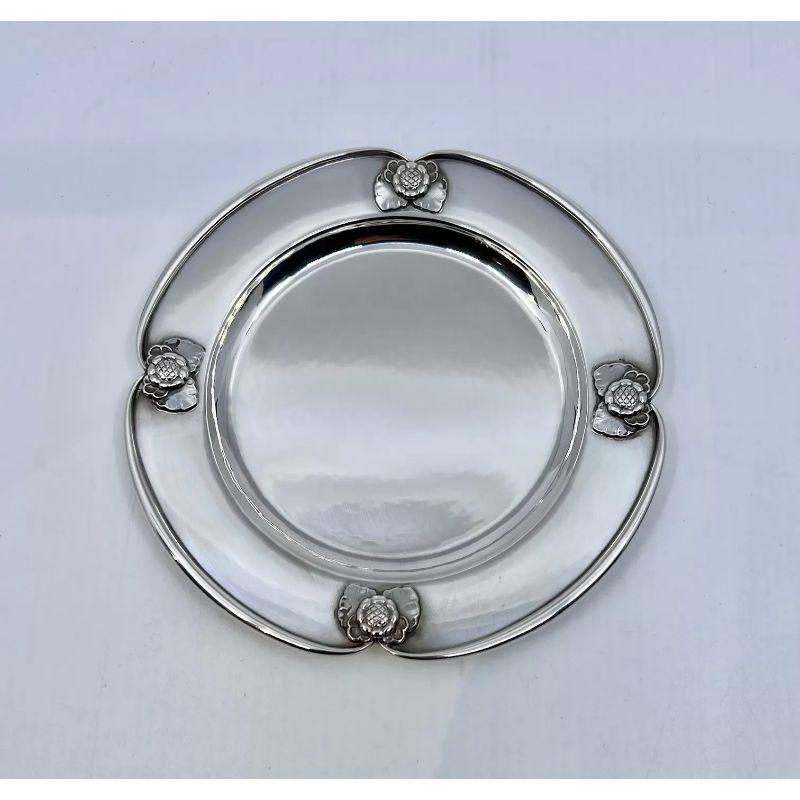 The rare Georg Jensen Bread plate or bottle coaster holder from circa 1927 is a sterling silver masterpiece. Design #491B showcases intricate floral motifs with visible hand-hammering. Its heavy, high-quality construction and curved edges
