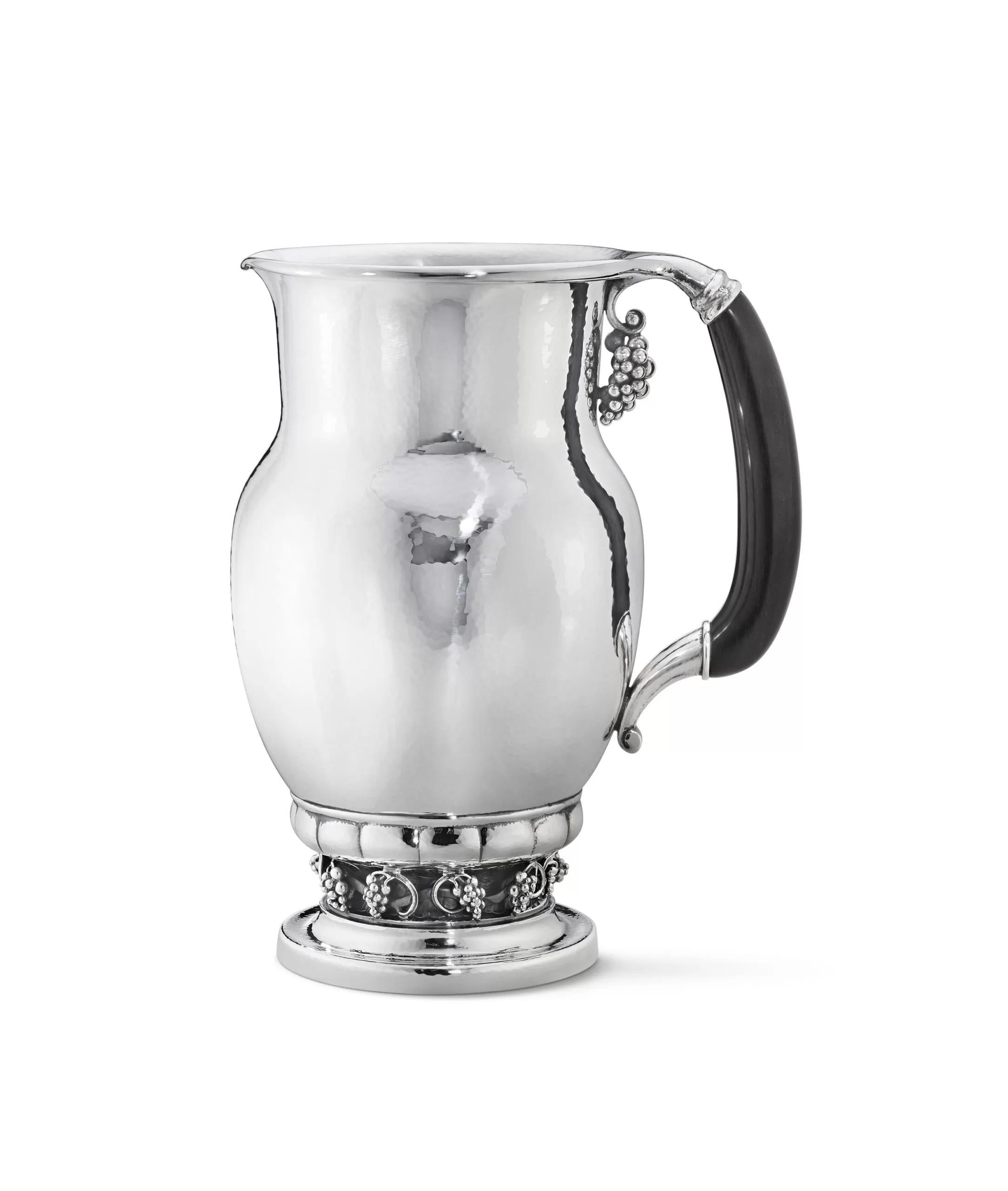 Esta jarra Georg Jensen de plata de ley, diseñada por Georg Jensen en 1925, es un impresionante ejemplo de la artesanía de principios del siglo XX. Con una base graciosamente redondeada, la jarra asciende hasta un racimo de uvas que rodea el cuerpo,