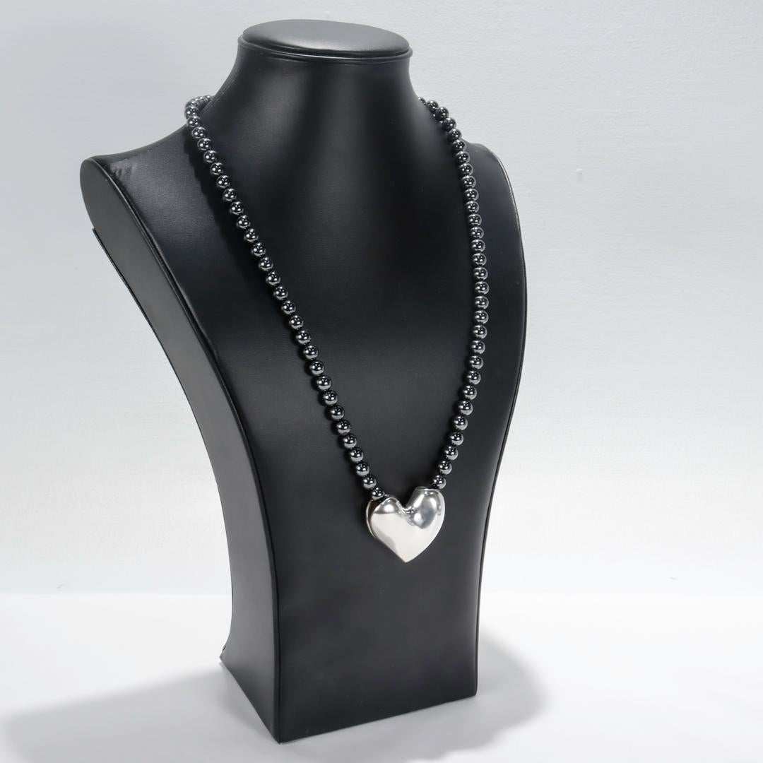 A fine sterling silver & hematite necklace.

By George Jensen.

Designed by Allan Schaff.

With a sterling silver pendant in the shape of a heart suspended from a strand of hematite beads. 

The hematite beads have a sterling lobster clasp.

Marked