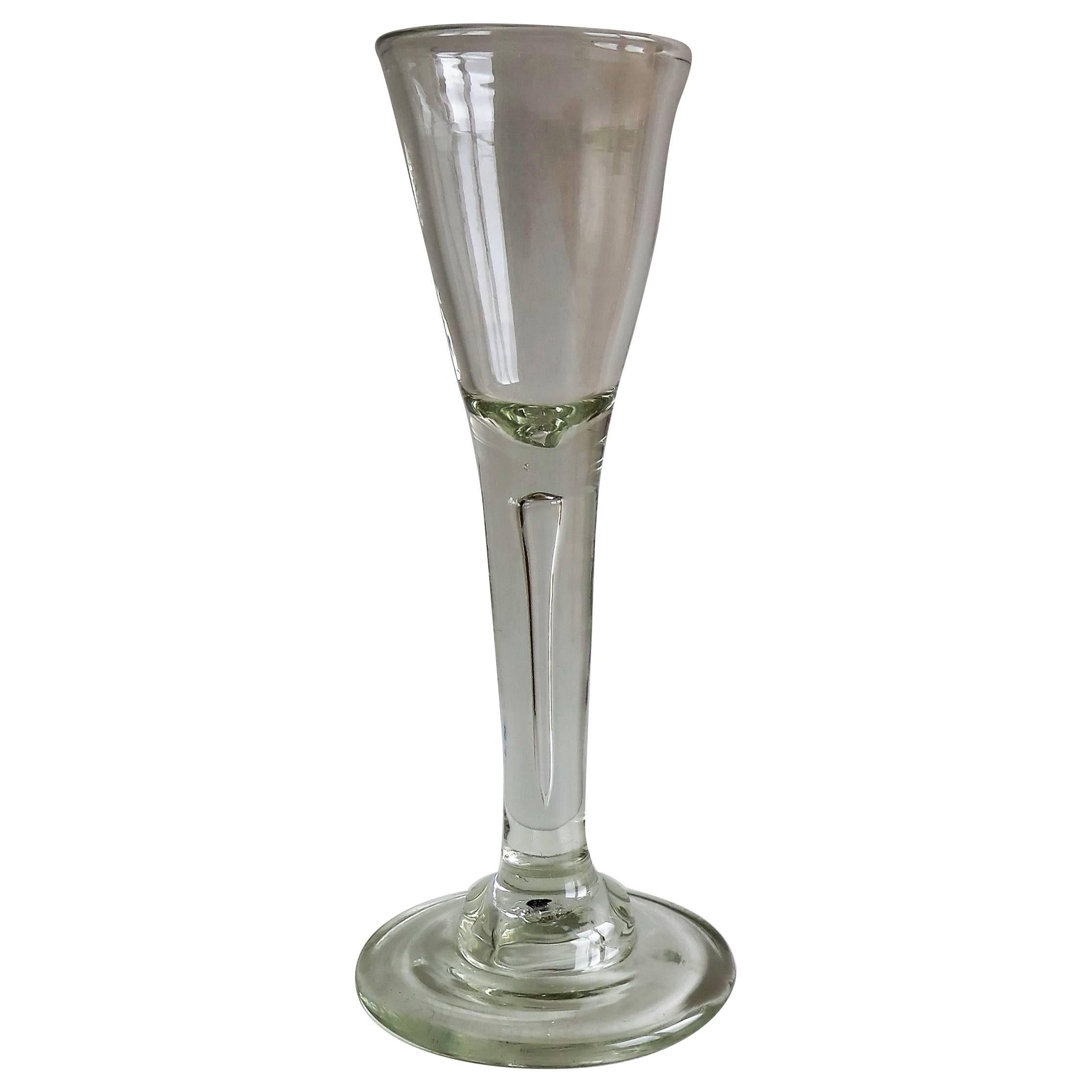 This is a tall mid-18th century, hand blown, wine glass with a drawn round funnel shaped bowl having a solid base. The bowl extends into a tapered solid drawn stem with a long tear (air bubble formed by the glass blower) through most of its length.