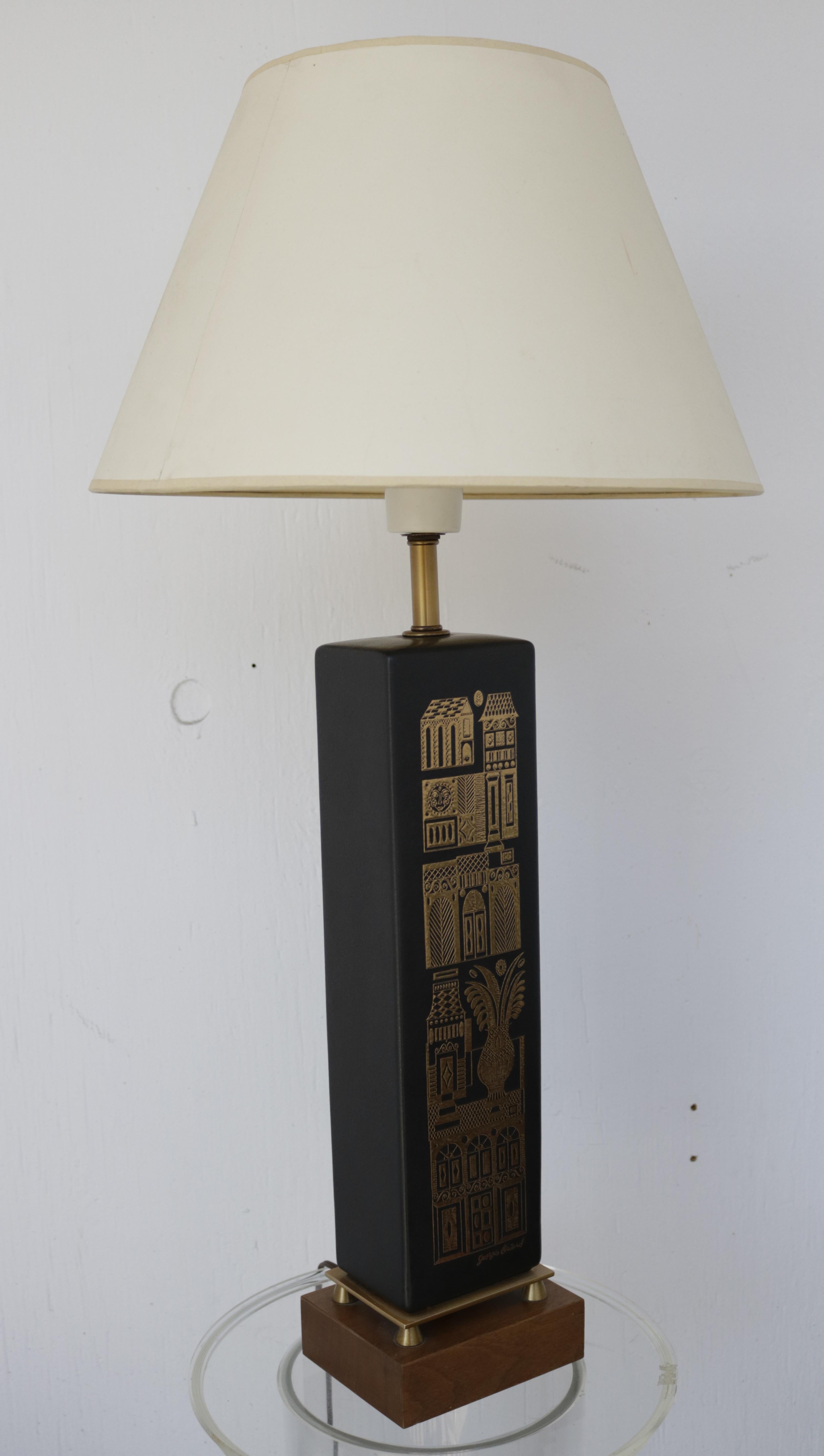 This stunning 1960s matt black ceramic accent lamp by Georges Briard features a gold embossed village scene and the signature of the artist. 
It stands on a brass stand and a walnut base. The off-white paper lampshade rests on a plastic holder that