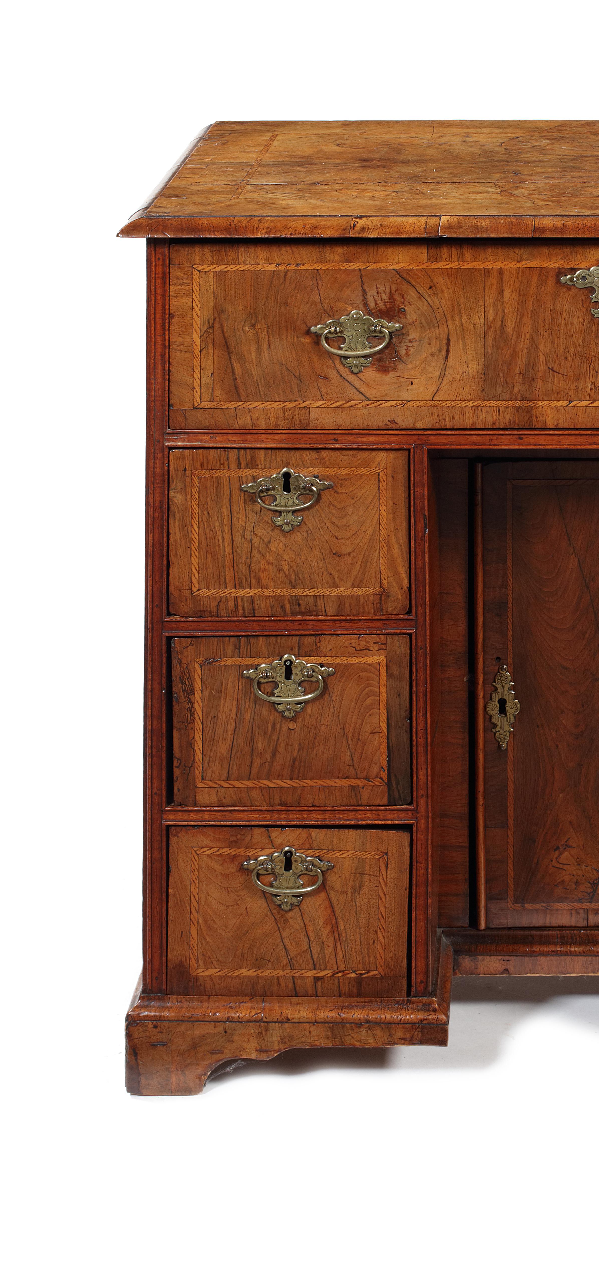 A George I walnut and ash banded kneehole desk
The quarter-veneered top above a frieze drawer and three short drawers to each side, flanking a kneehole cupboard door enclosing a shelf; on short bracket feet, 80cm wide, 46cm deep, 78cm high.