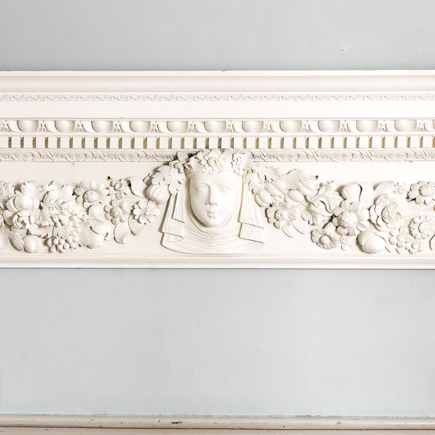 A George II carved pinewood overdoor, in the manner of William Kent, the deep cornice with ovolo, egg and dart and dentil mouldings above a frieze centred by female mask flanked by swags of fruit and flowers, with elongated scrolled brackets to