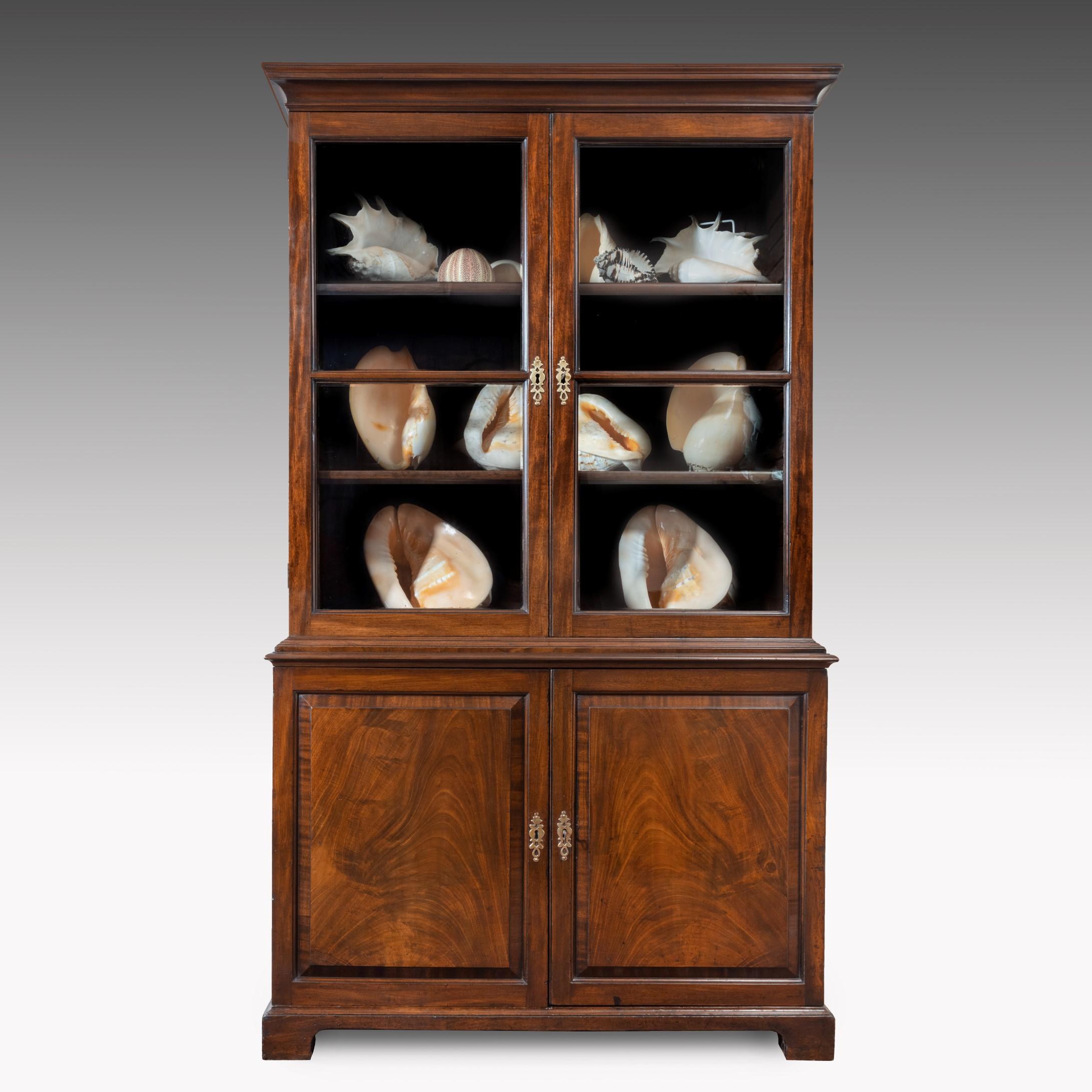 A handsome George II mahogany cupboard base bookcase; the cavetto moulded cornice above a pair of glazed doors which open to reveal adjustable shelves; below are a pair of fielded panel doors veneered in beautifully figured Cuban mahogany and open