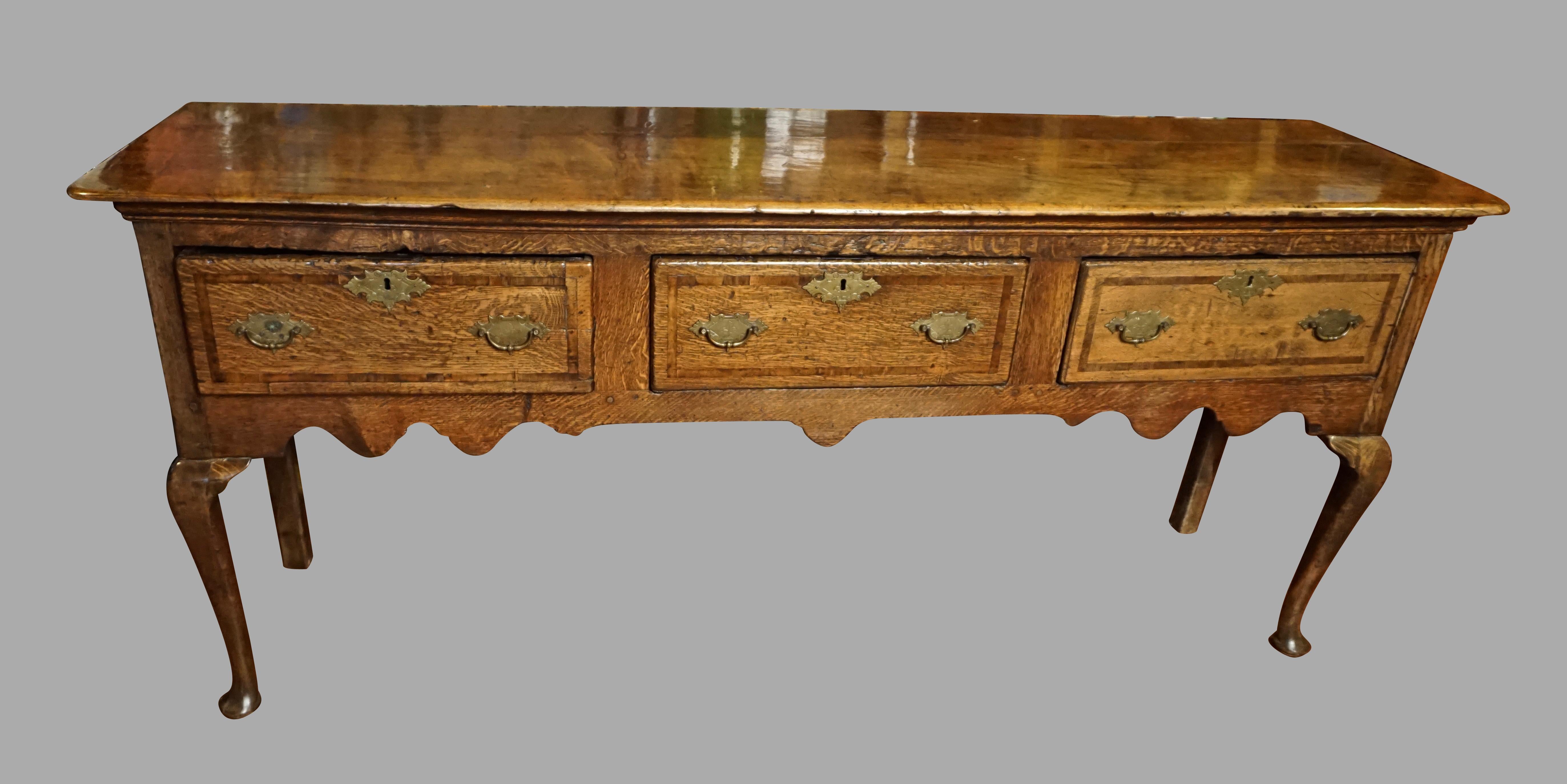A good quality English oak dresser base, the molded overhanging top above 3 mahogany inlaid crossbanded drawers, the front with a wavy apron, supported on front cabriole legs ending in pad feet. 
Pegged construction. Replaced hardware. Circa 1760.