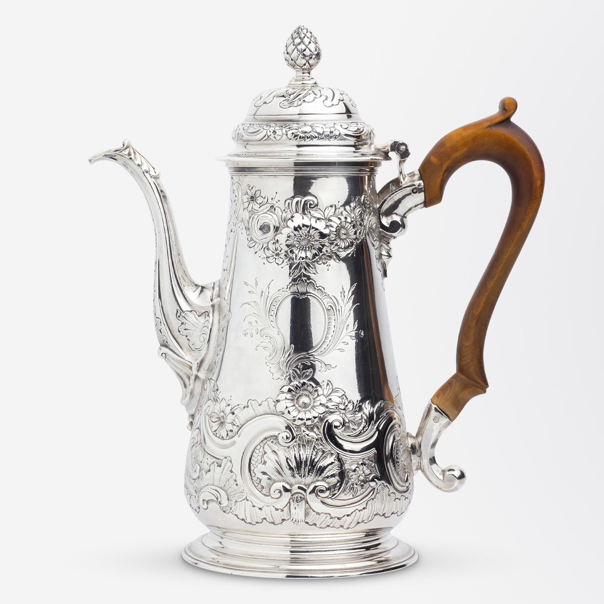 This tall George II period sterling silver coffee pot is decorated with elegant floral repousse details and features a blonde timber handle. To the side of the coffee pot there is a blank cartouche which remains free of a heraldic crest or a