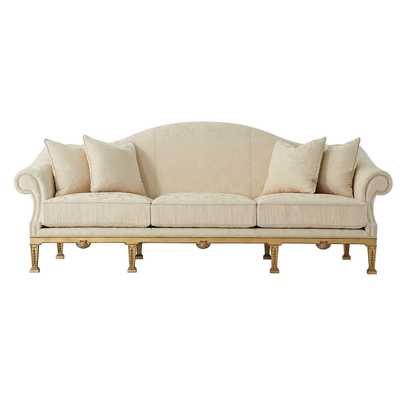A George II style finely upholstered camel back sofa, the tightly upholstered back above three loose seat cushions with four throw pillows enclosed by out scrolled arms, on a hand-leafed gilt base carved in mahogany, with shell details and square