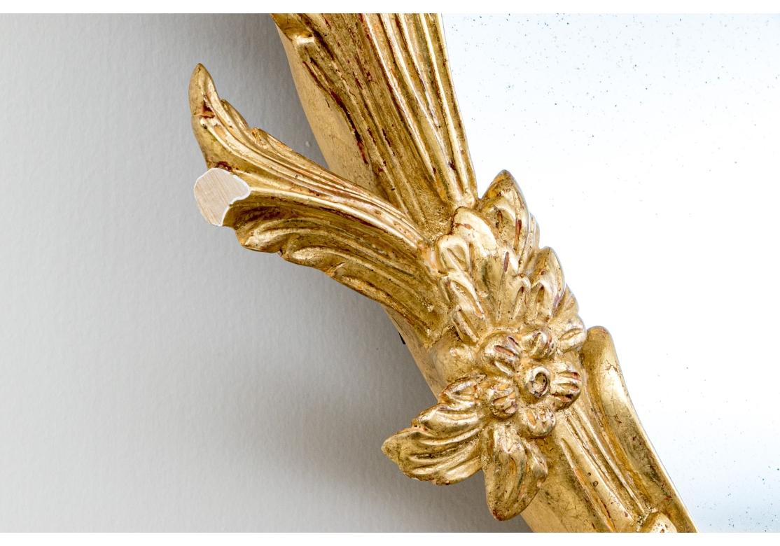 A refined oval mirror in an exceptional George II style gilt frame with an ornate palmier-scrolled pediment with foliate flourishes complimenting a leafy wreath design around the perimeter, accented with crimped ribbon motifs at the