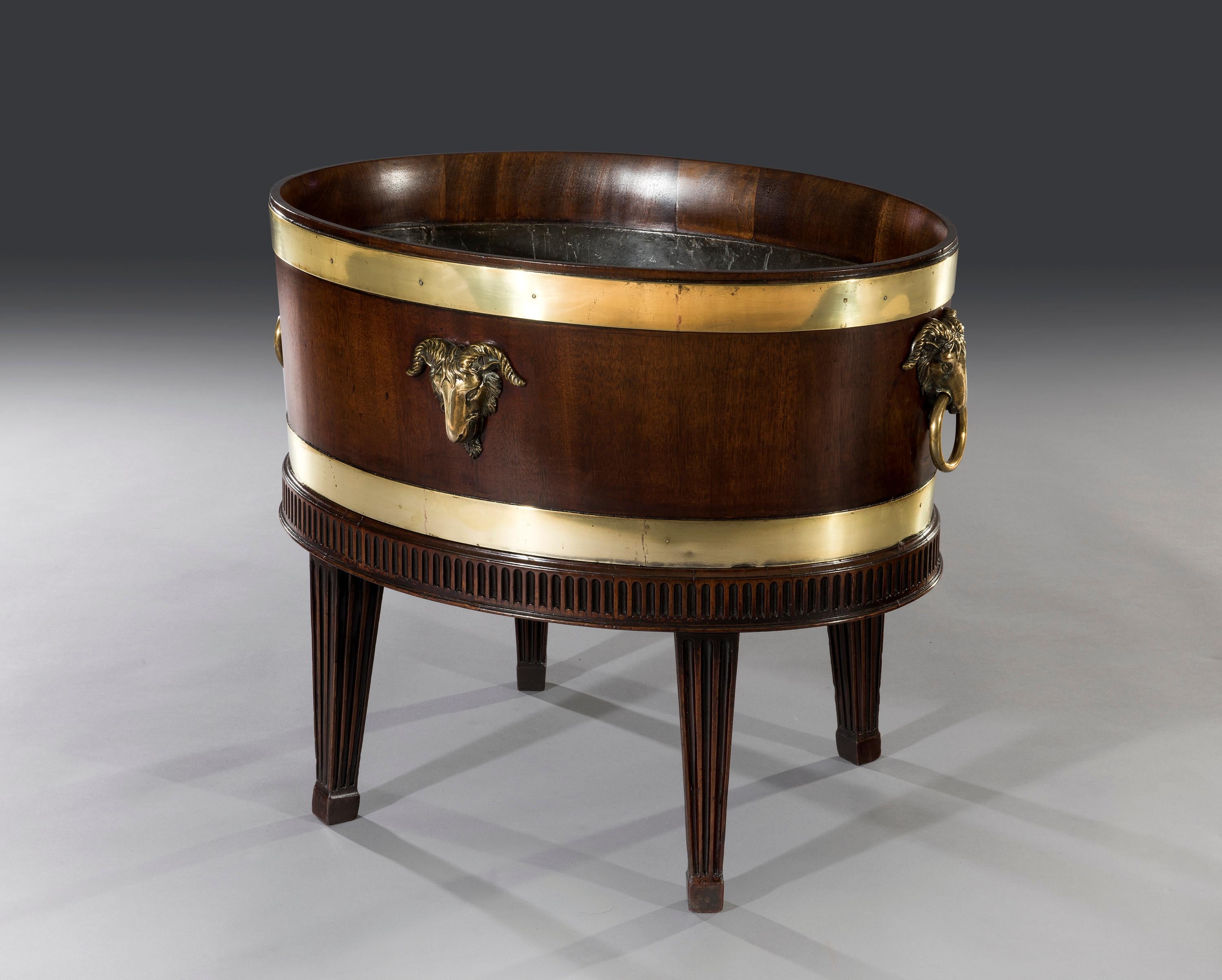 The mahogany oval shaped lead lined wine cooler has brass bands and is decorated with four ram's head brass fittings. The detachable mahogany base with a fluted frieze and fluted square tapered legs terminates on block feet.

The cooler has a dual