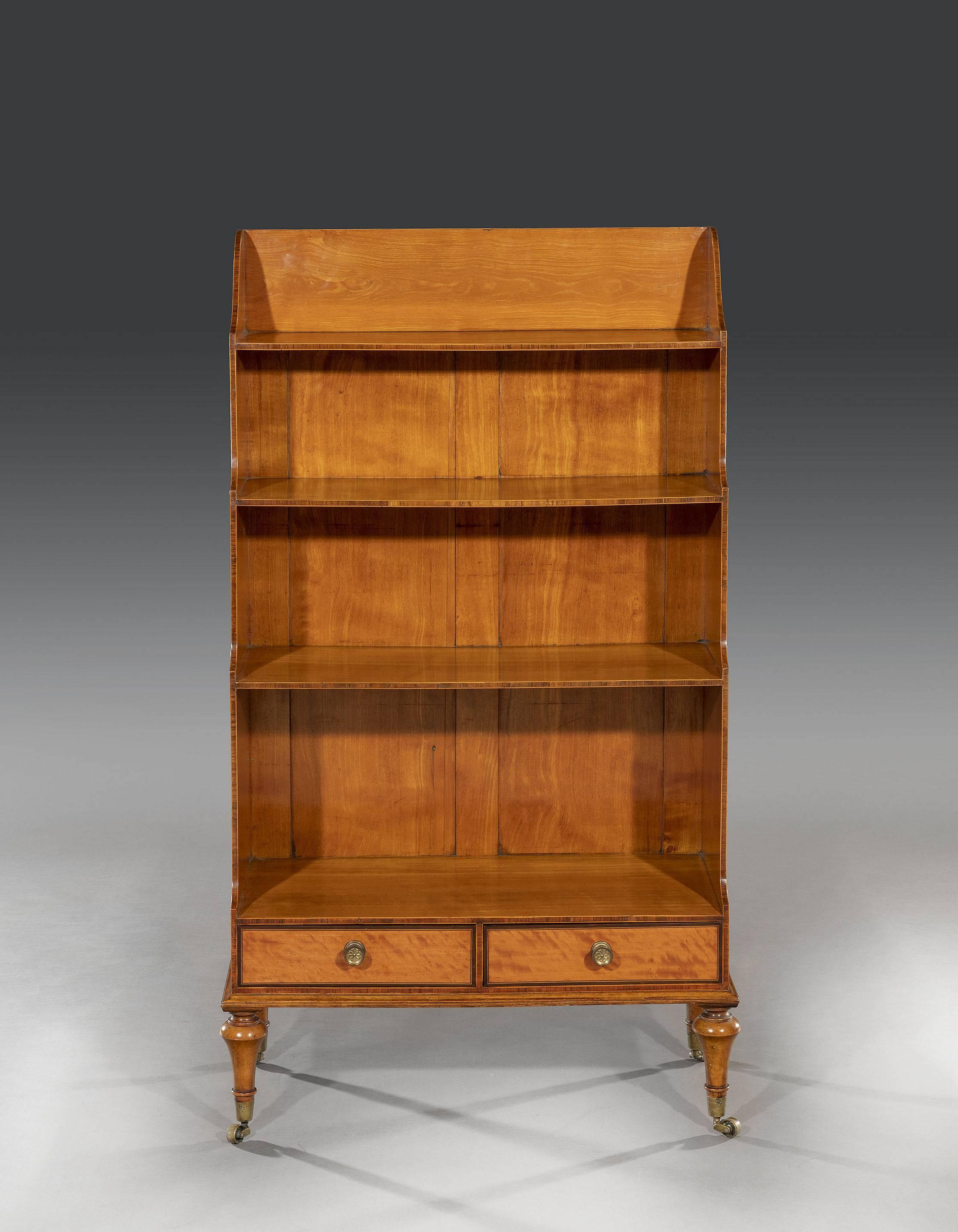 An elegant satinwood waterfall bookcase with veneered backboards and solid sides with kingwood cross banding through out. The graduated shelves sit above two drawers that retain the original brass handles and purple heart cross banding above solid