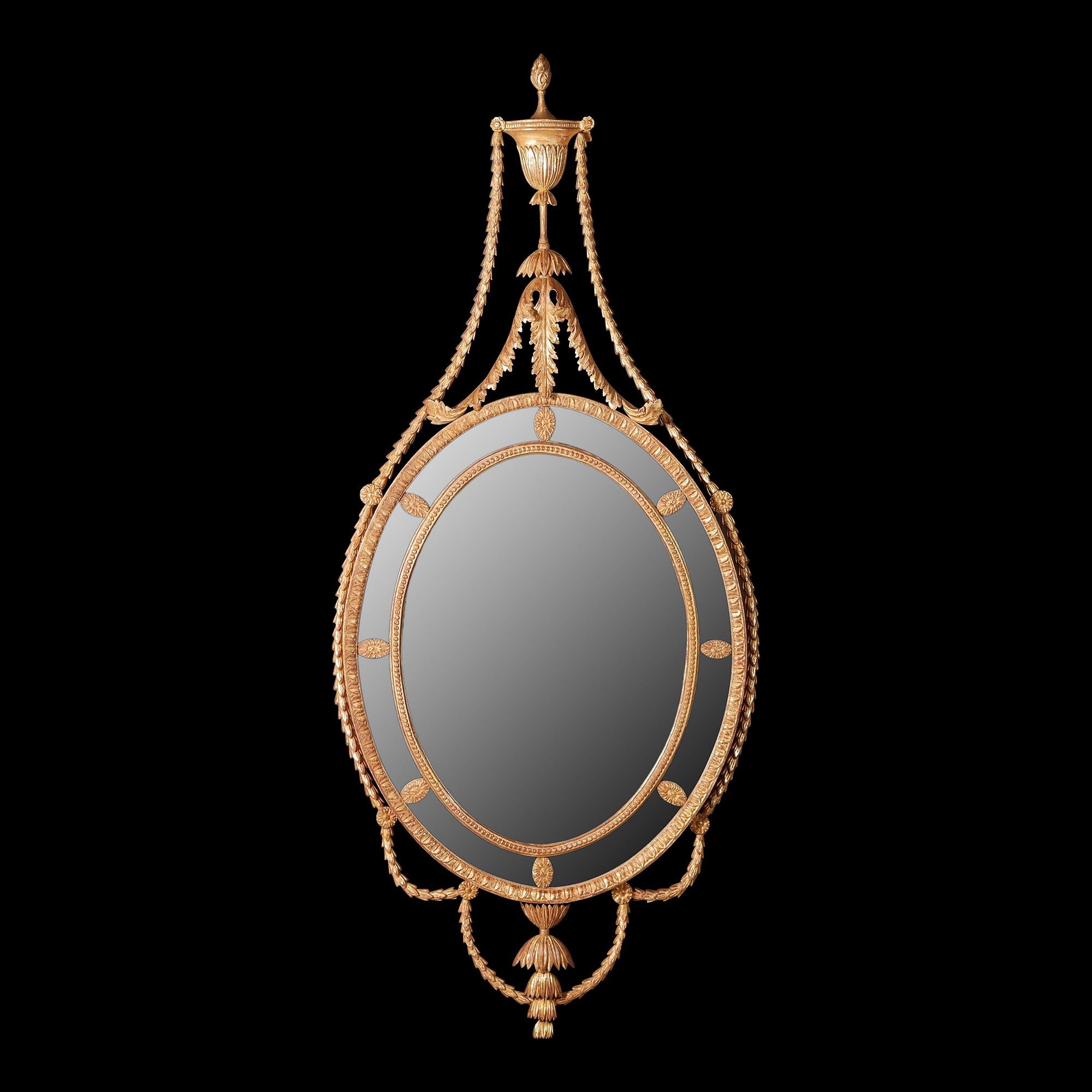 A carved oval mirror of Adam design with leaf carved outer frame and bead carved inner frame, the surrounding border glasses are punctuated by oval flower-head paterae. The cresting formed of a classical urn. The flame finial is raised upon a
