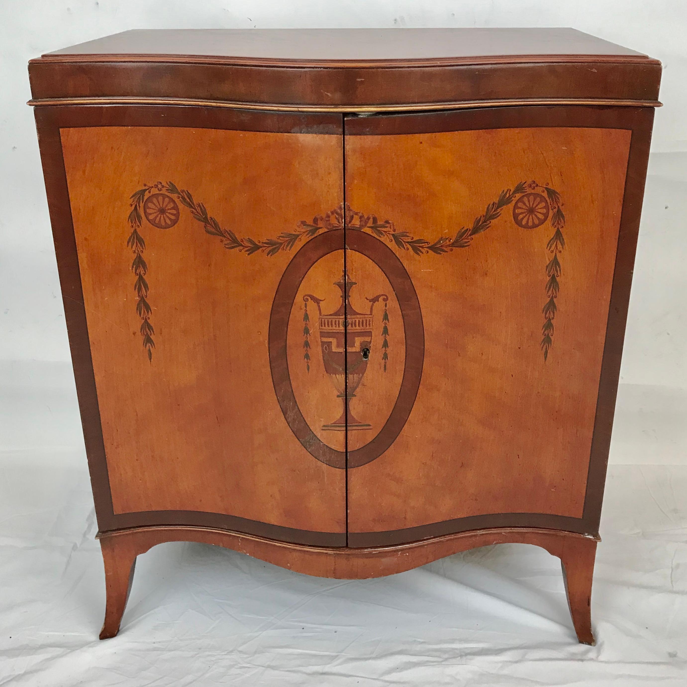 This handsome serpentine front satinwood cabinet features painted Neo-Classical style motifs, including bellflower swags, rosettes, and a lidded urn. 