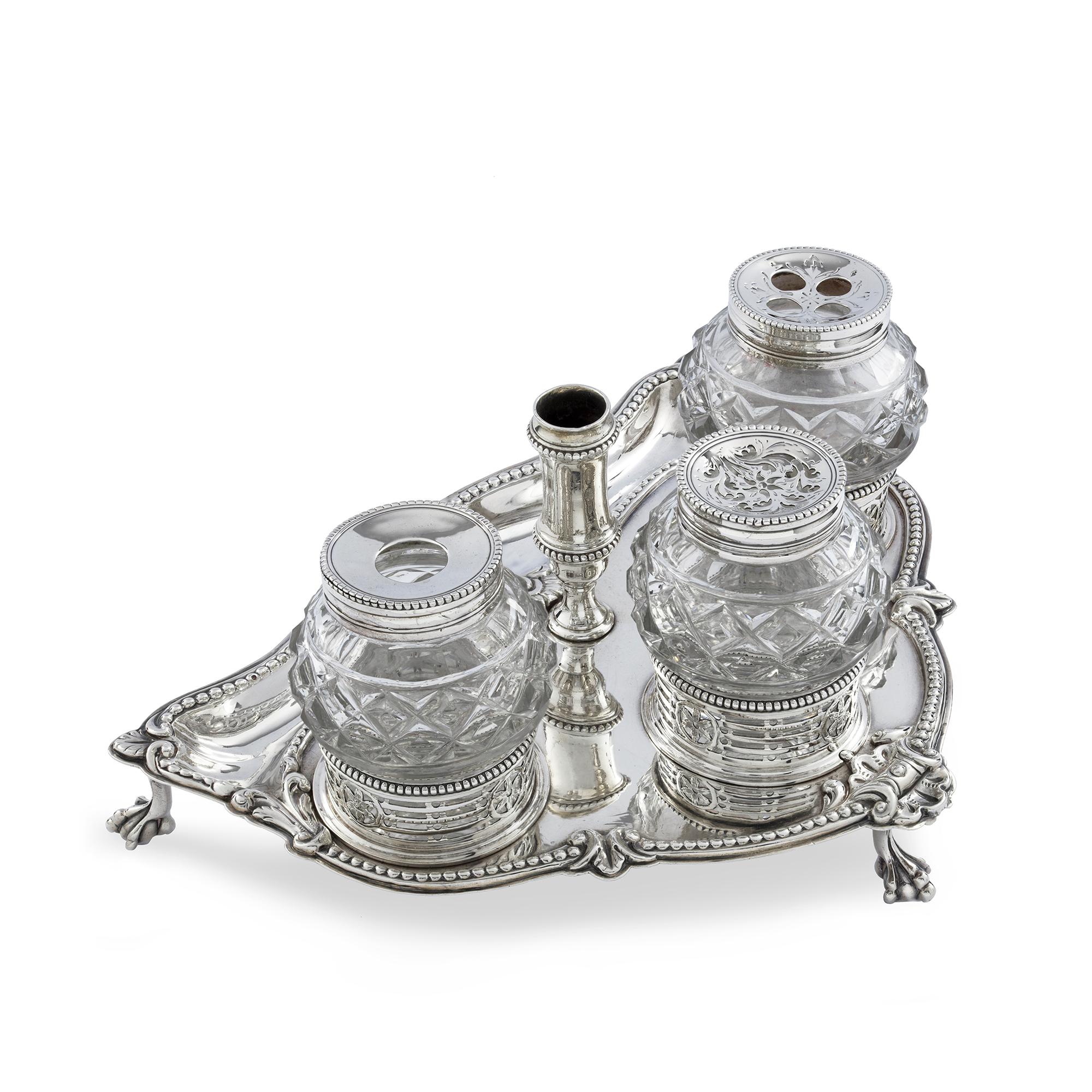 An Antique silver ink stand, comprising of three cut glass pots, silver removable lids, with beaded and moulded rim, placed within circular pierced borders with floral motifs, balustrade shaped pen holder, silver shield  tray with beaded rim and