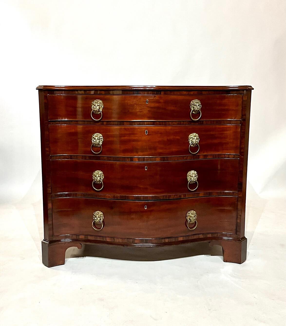 A fine circa 1770 George III period Chippendale style commode or chest of four drawers of serpentine form having mahogany case with later circa 1810 Regency period 'redecorating' of Gonçalo alves cross-banding and finely-cast gilt-brass lion's head