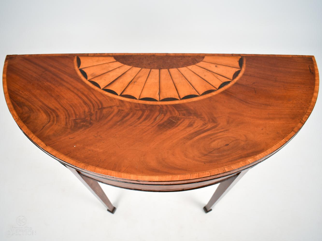 George III demi lune tea table with Sheraton design. Circa 1780 with deep dark surface colour inside the fold out top, mahogany and satinwood, on four tapered legs.