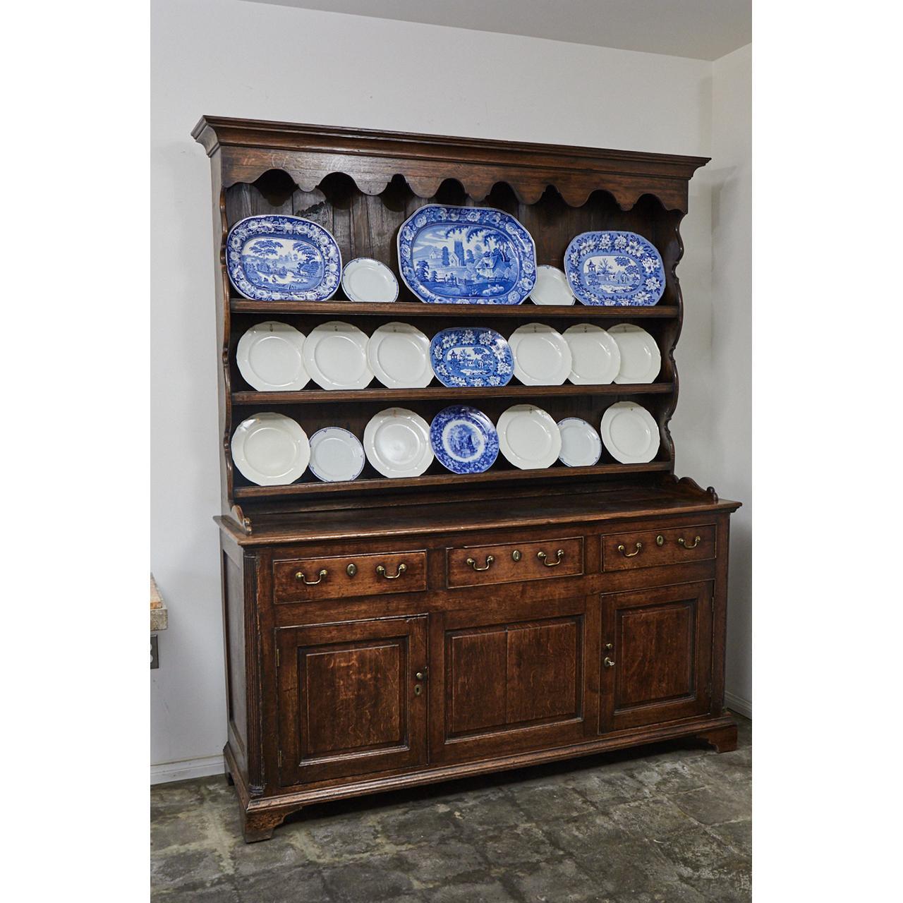 This English oak dresser has an 18th century base with a 19th century top. The piece has three shelves over three drawers and two doors. Each shelf has a plate holder. The top has carved decorative elements on the top and sides. The base has
