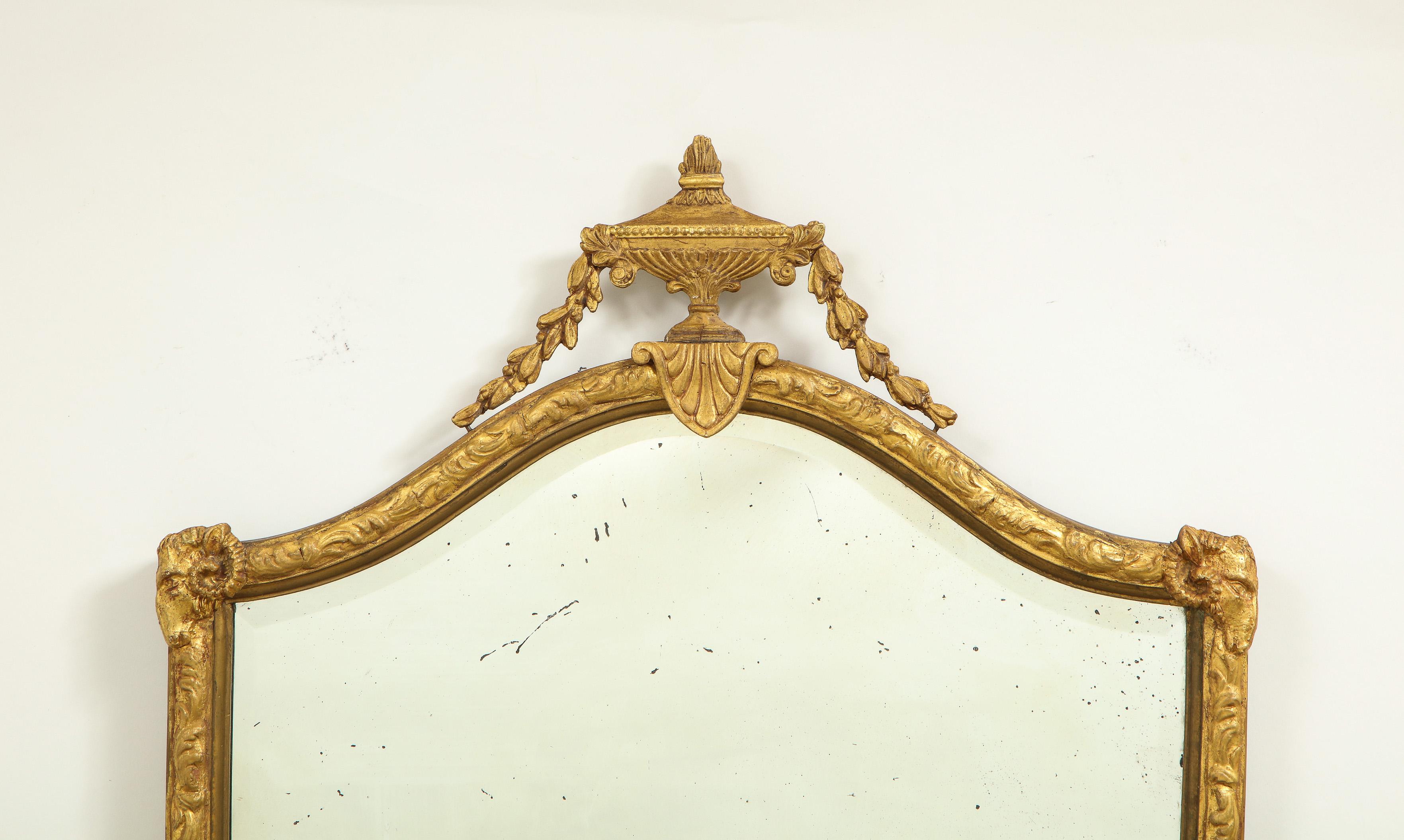 The shield-shaped mirrored plate set within a conforming gilt gesso surround enriched with foliate decoration; surmounted by a bell garland-festooned classical urn resting on a paterae lambrequin.