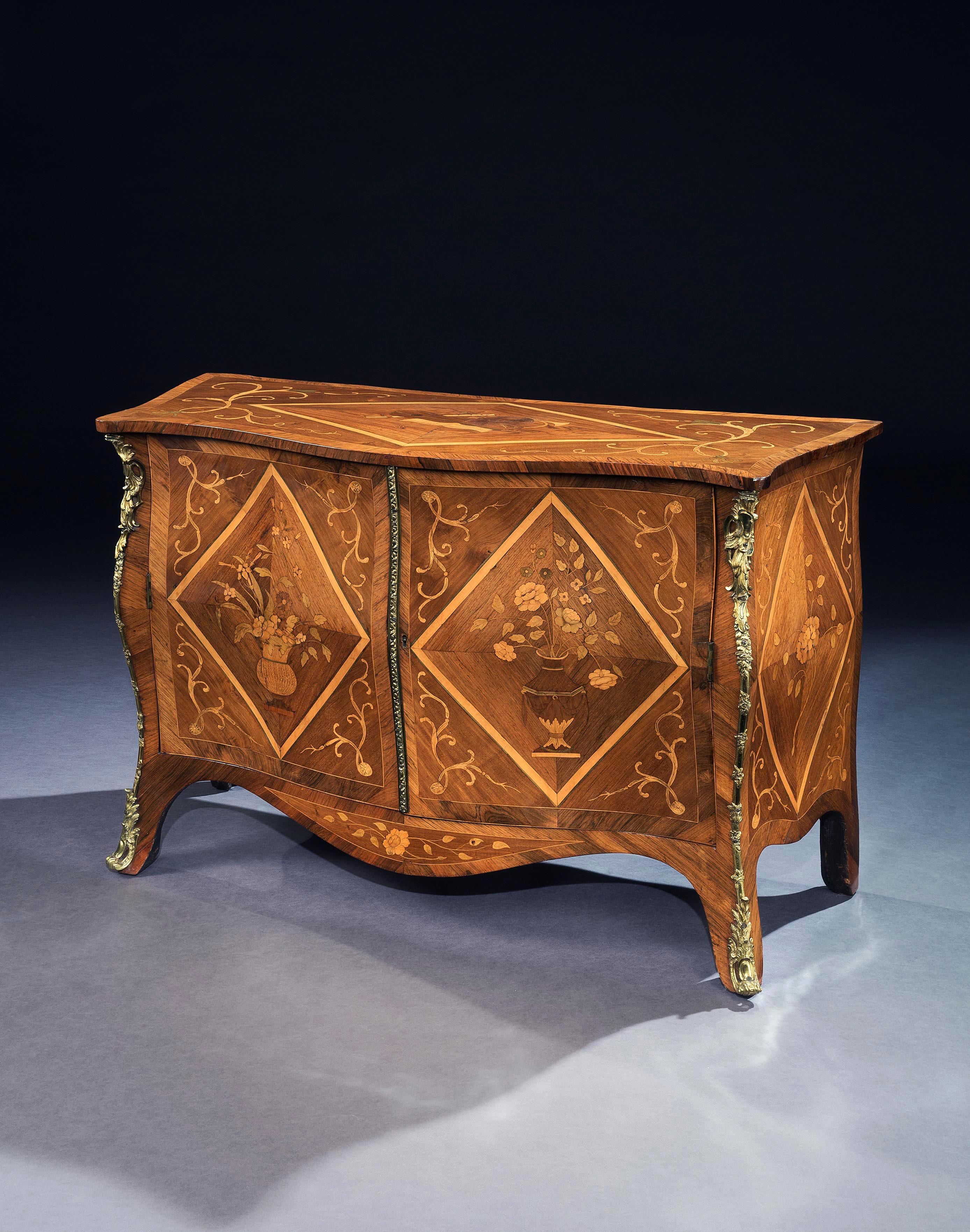 A George III bombé commode veneered in rosewood and hardwood, the cross banded top inlaid with scrolls and a central diamond form enclosing a marquetry panel depicting a musical score and various musical instruments.
The lower part with a pair of