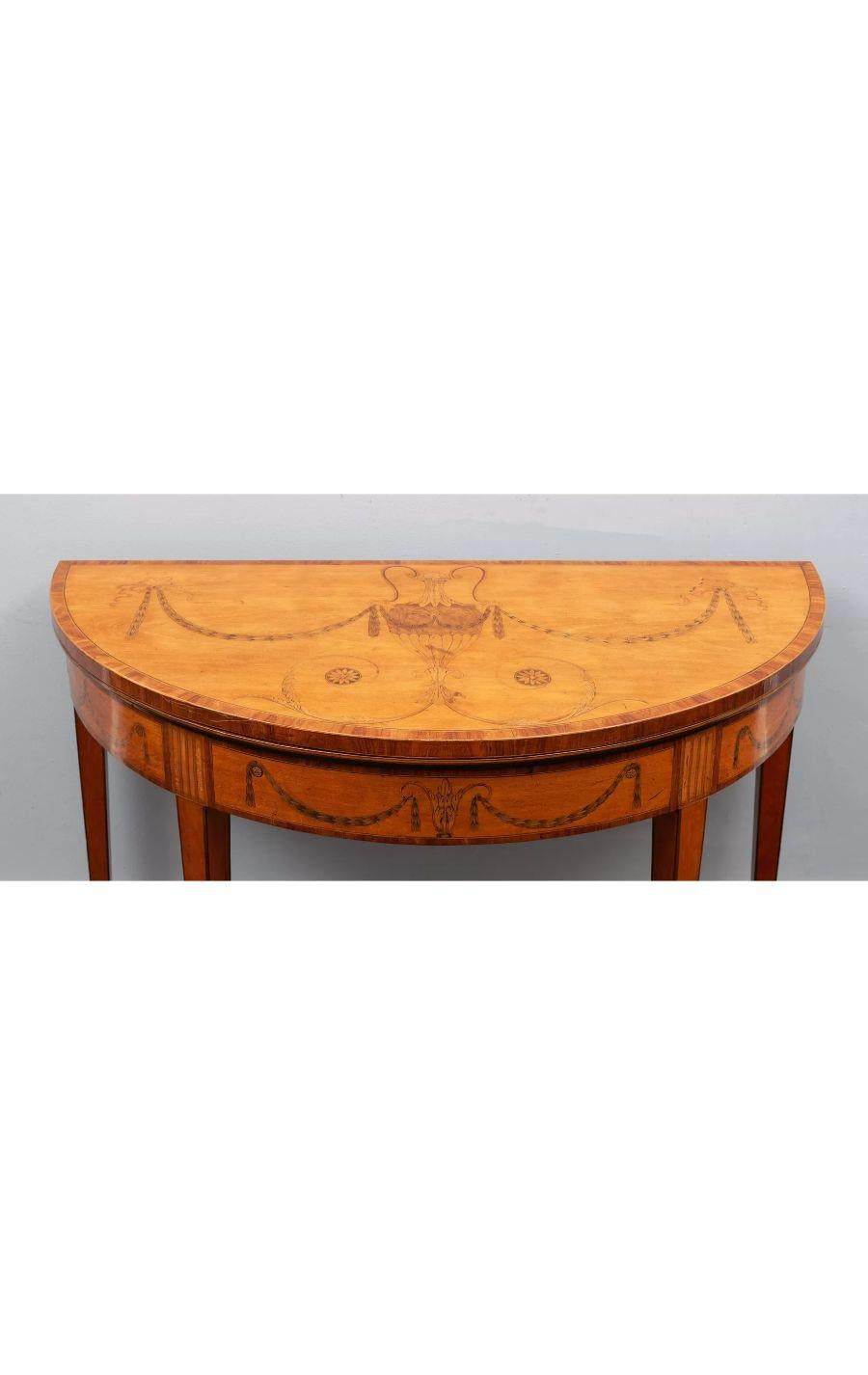 Ein Demi-Lune-Tisch mit Intarsien aus Satinholz nach William Moore, George III.

Neoklassizistischer Stil mit besonders schönen Intarsien und Einlegearbeiten aus exotischen Hölzern.

Die sich verjüngenden Stützen enden in Messingrollen.

CIRCA