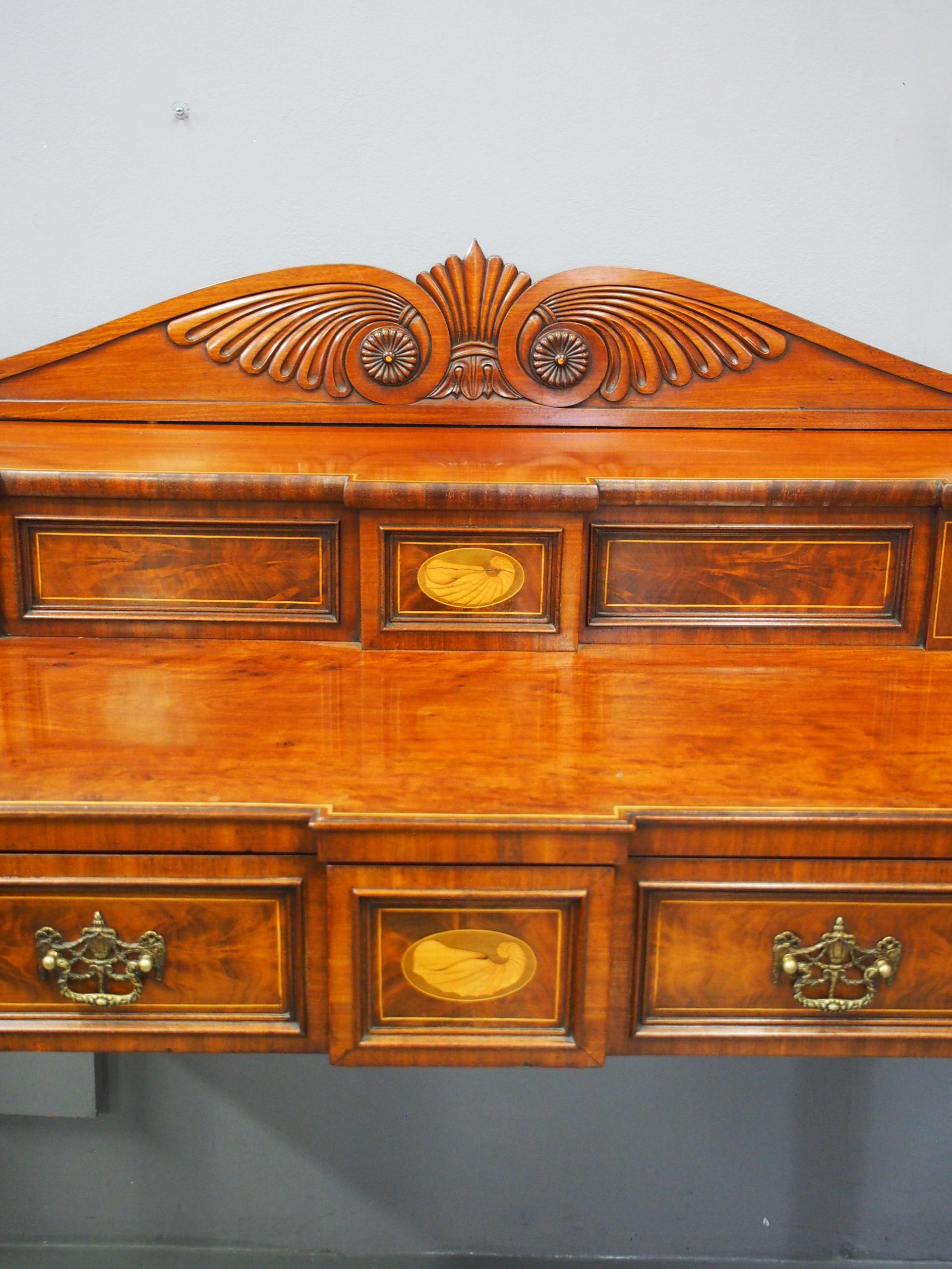 George III inlaid Scottish mahogany sideboard, circa 1800. The stage back with boxwood stringing and shell and urn inlaid panels with sliding doors surmounted by a shaped, acanthus-carved back. The breakfront top in plum pudding mahogany is above