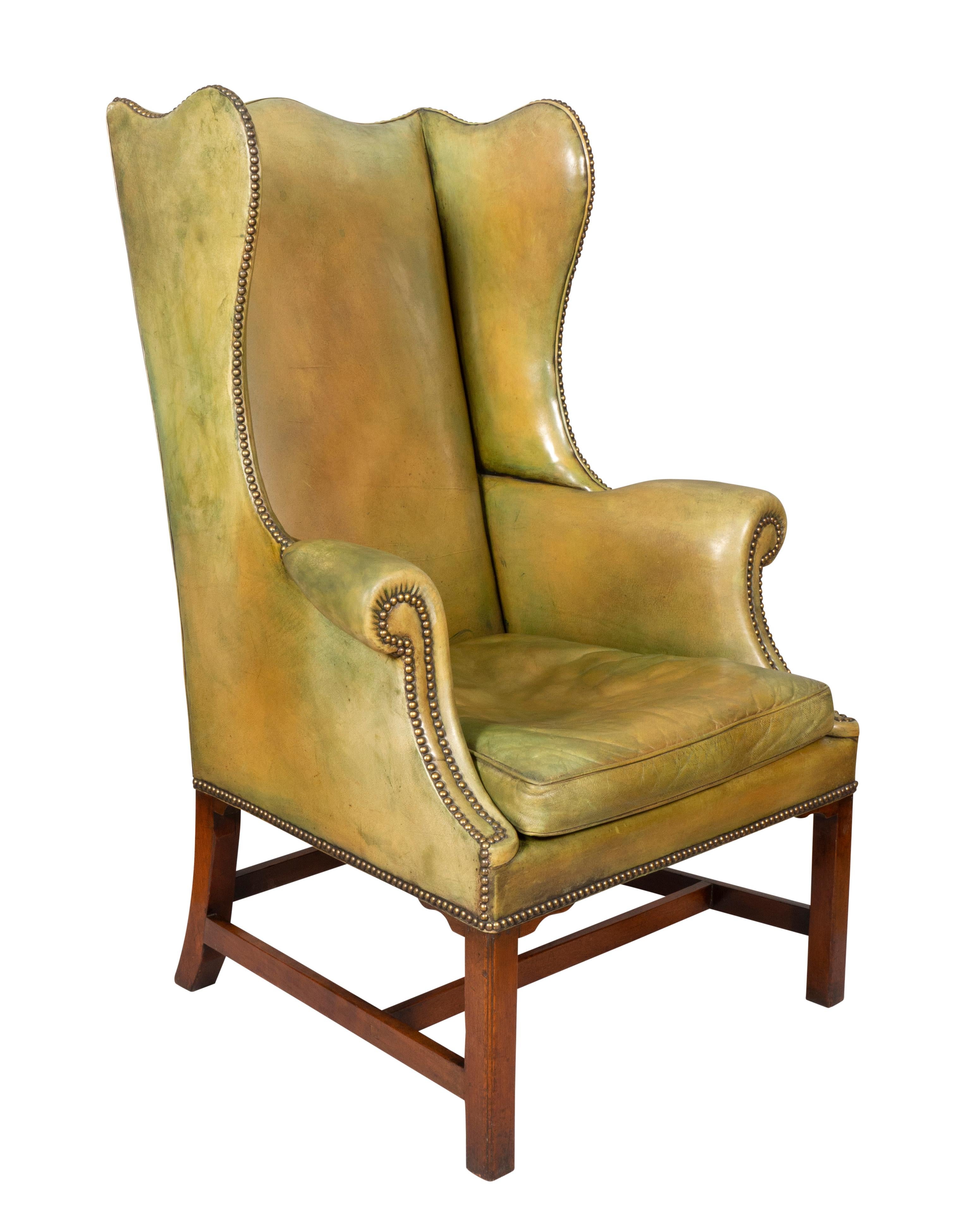 With green leather faded into some yellow tones. Typical form with wing back and scrolled arms with brass tacks and raised on square section legs and double H form stretchers.