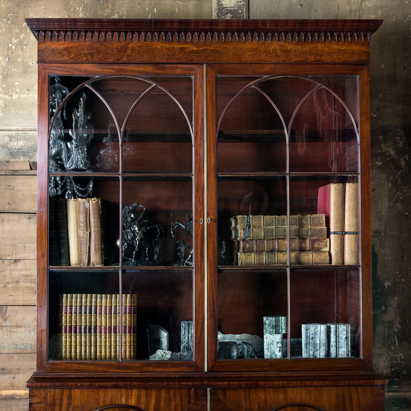 A late George III Mahogany bookcase, the moulded cornice above frieze hung with gothic pendants, the gothic glazed doors with astragal bars moulded as cluster columns, above base with oval panels to the doors enclosing an arrangement of drawers
