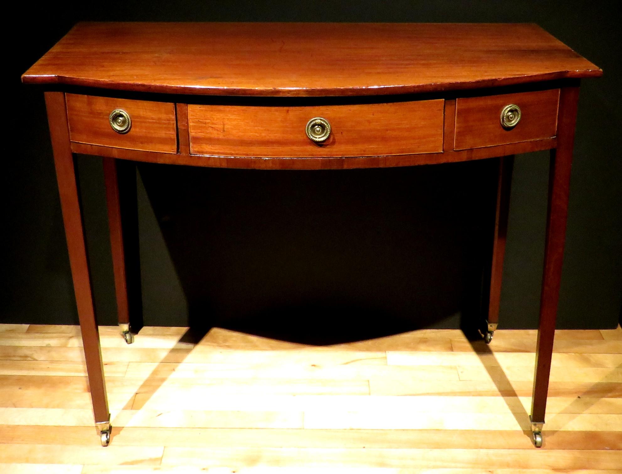 A very handsome early 19th century Georgian bow-fronted writing table / sofa table in mahogany, showing a bow-fronted overhanging top above a conforming frieze fitted with three drawers having period brass ring pulls and inlaid boxwood stringing,