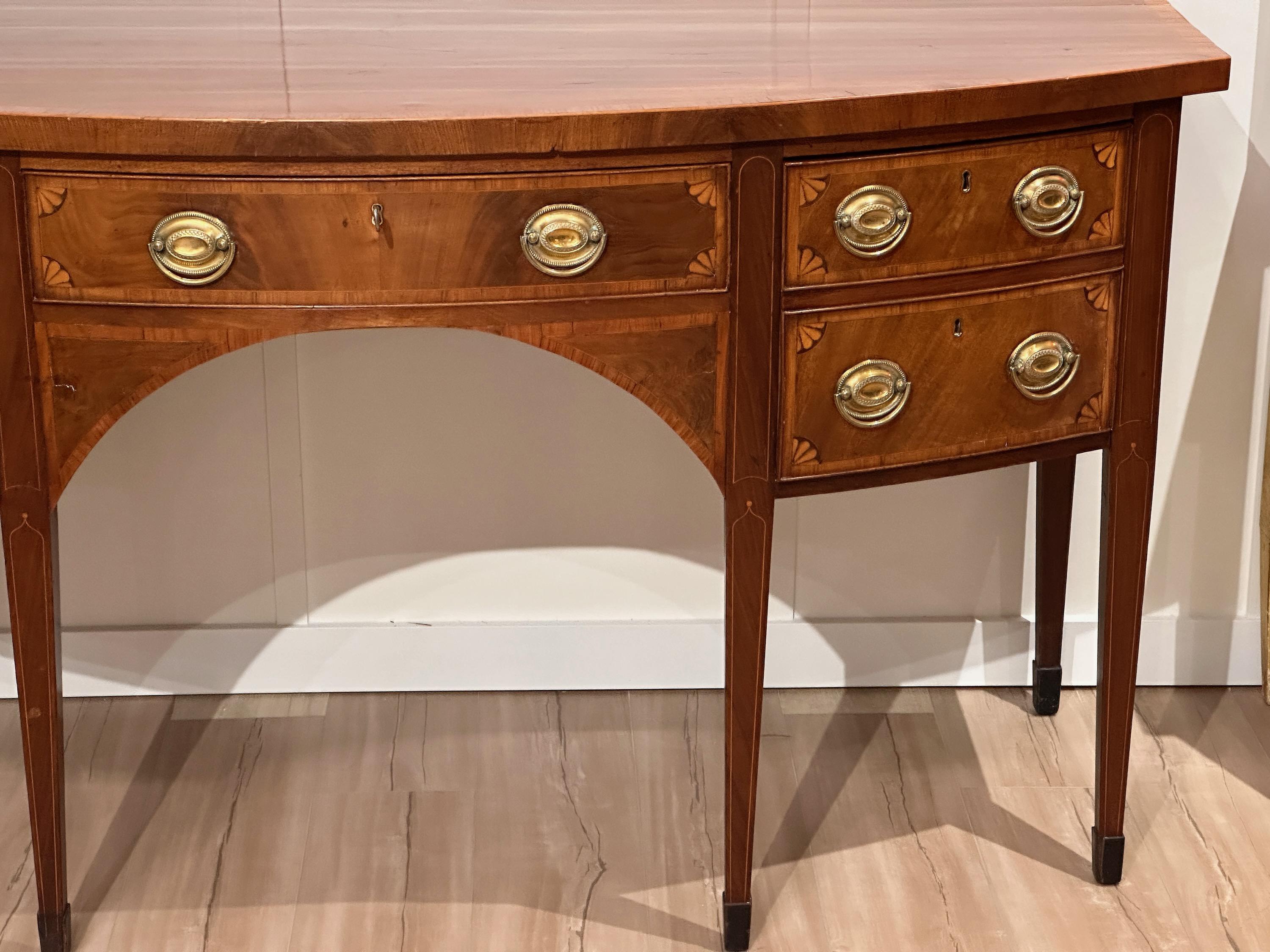 An English George III bowfront sideboard in mahogany with corner fan satinwood inlay and original oval gilt brass pulls, a central frieze drawer, flanked on the left with double drawers and on the right with a deep bottle drawer, all supported by