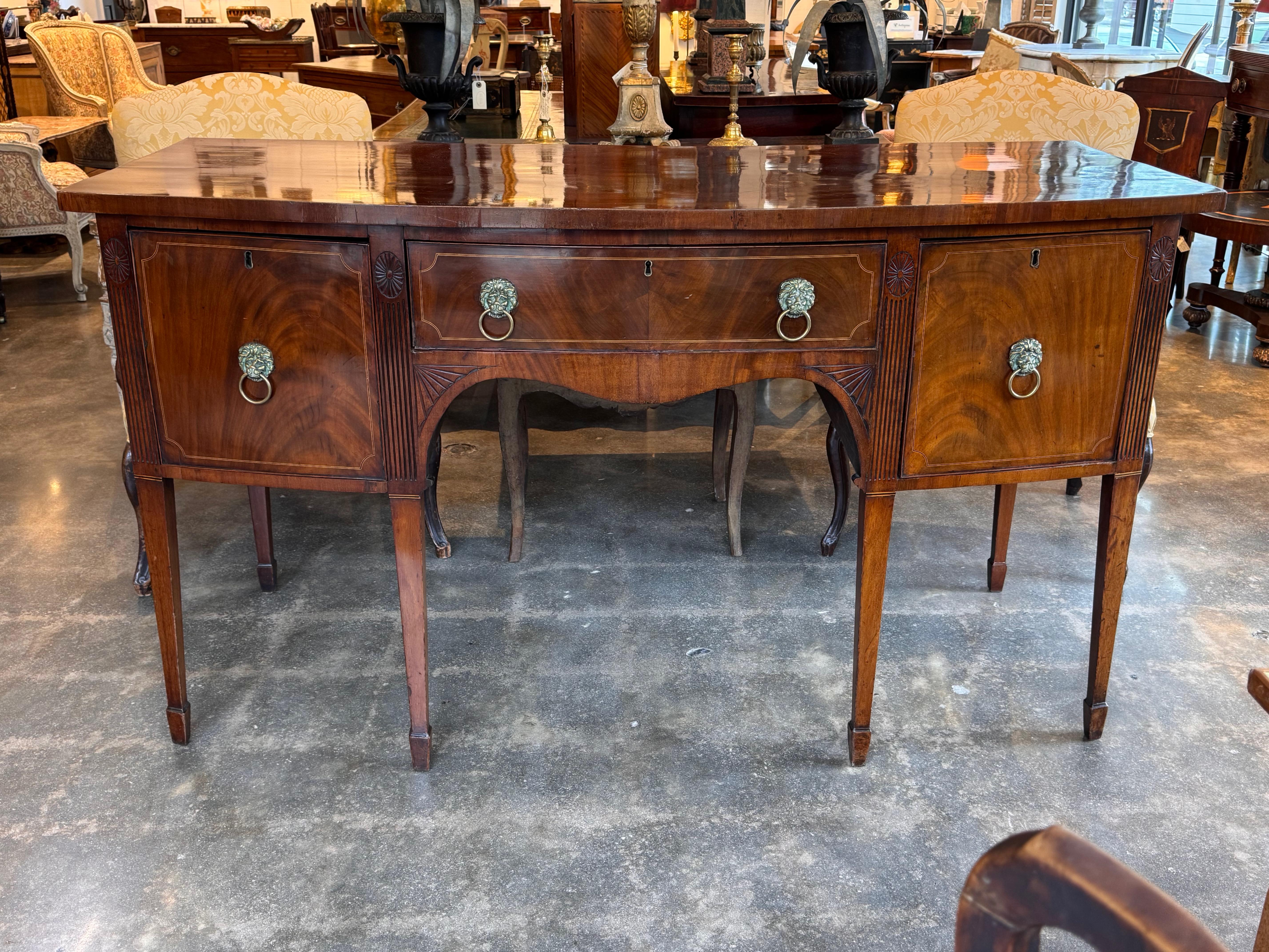 This George III period sideboard is crafted from richly figured mahogany and features a graceful bowfront form. The central drawer is flanked by two cupboard doors, all detailed with inlaid borders and original lion mask brass pulls. Reeded and