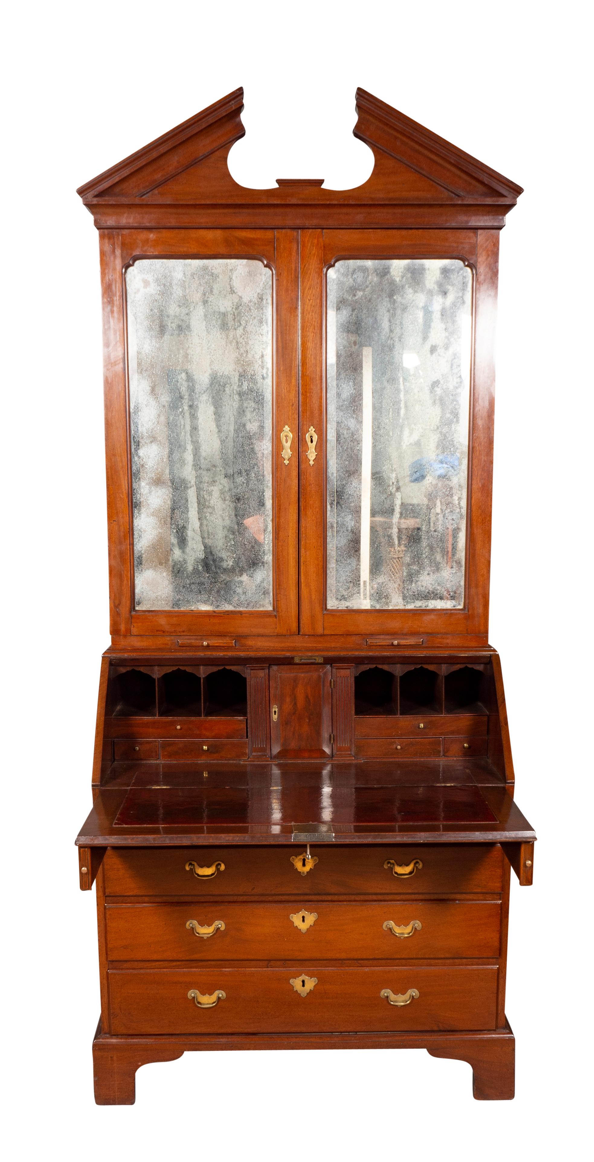 With architectural broken pediment cornice over a pair of mirror doors opening to a fitted interior, the base with hinged slant lid with red leather surface opening to a fitted interior over four graduated drawers and ending on bracket feet.