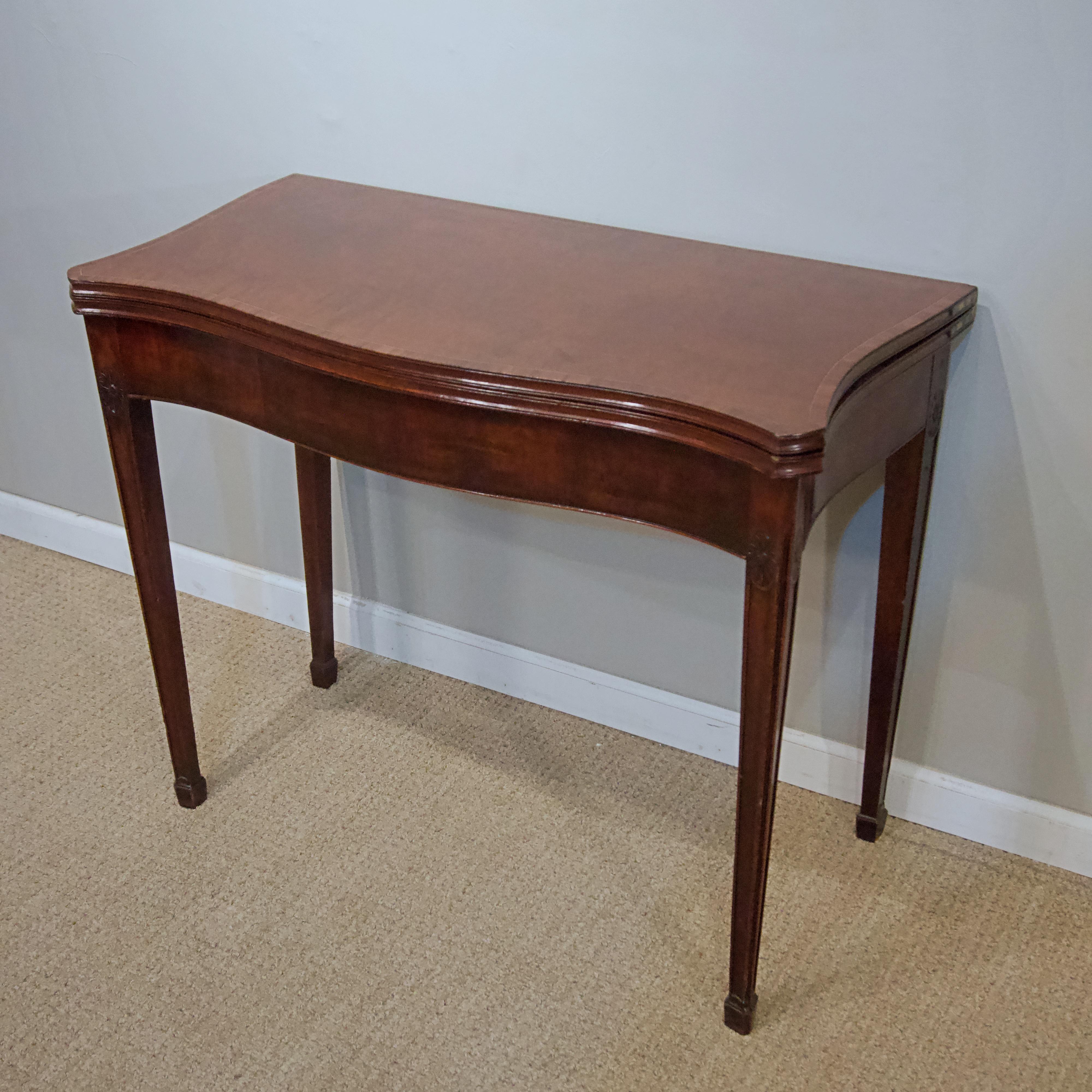 Mahogany card table. 
The serpentine-fronted top with canted corners opening to a baize-lined gaming surface 
above the square tapering canted legs carved with a oval paterae.

In the Adams taste; Dimensions- width 36”, depth 17 1/2”, height