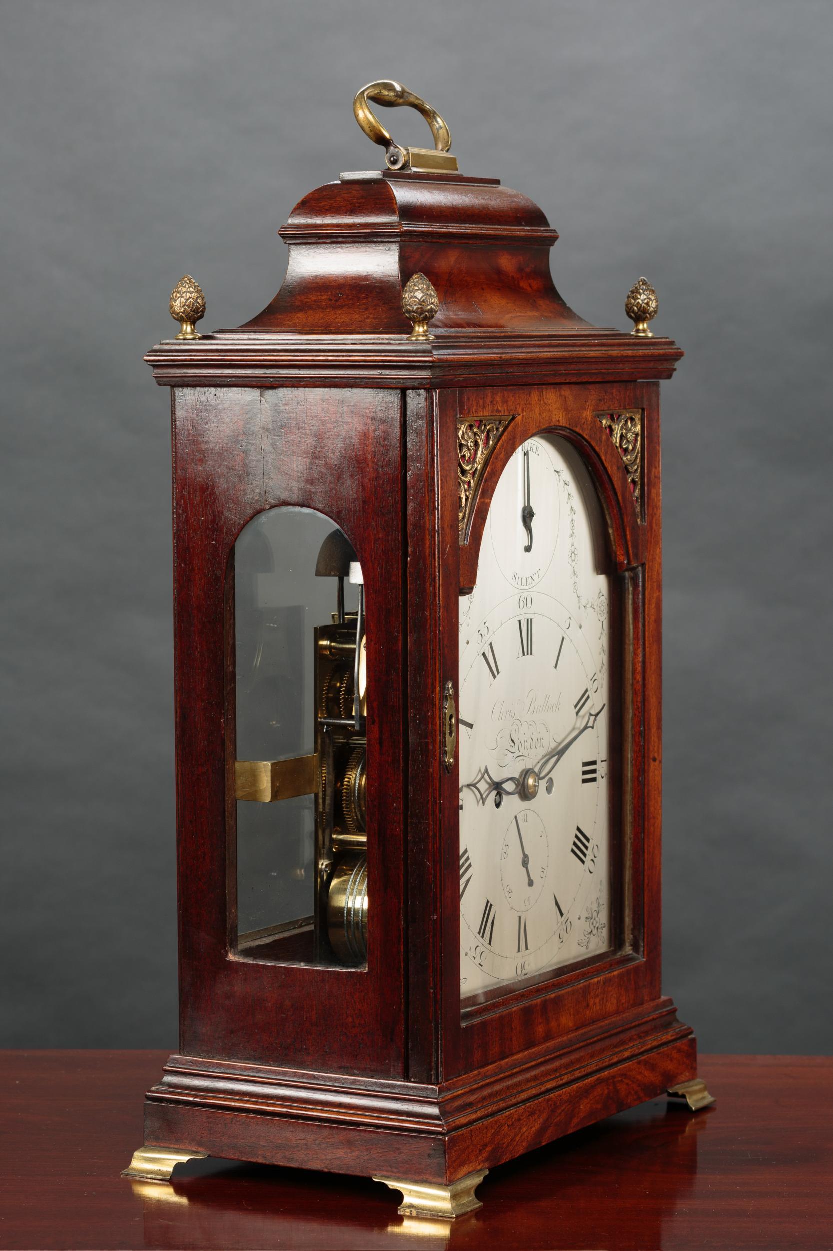 George III bracket clock by Christopher Bullock, London

Finely figured mahogany bell top case with four ‘pineapple’ finials surmounted by a hinged carrying handle standing on a stepped, raised plinth and resting on four outswept brass bracket