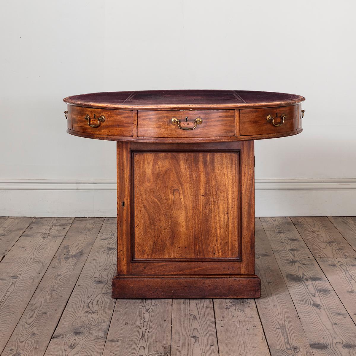 A George III Mahogany Drum Table With blind tooled red Moroccan leather panelled top, each triangular and curve fronted drawer labelled alphabetically, and with gilt lacquer swan neck handles of pleasing patina, the box base with ample cabinet space