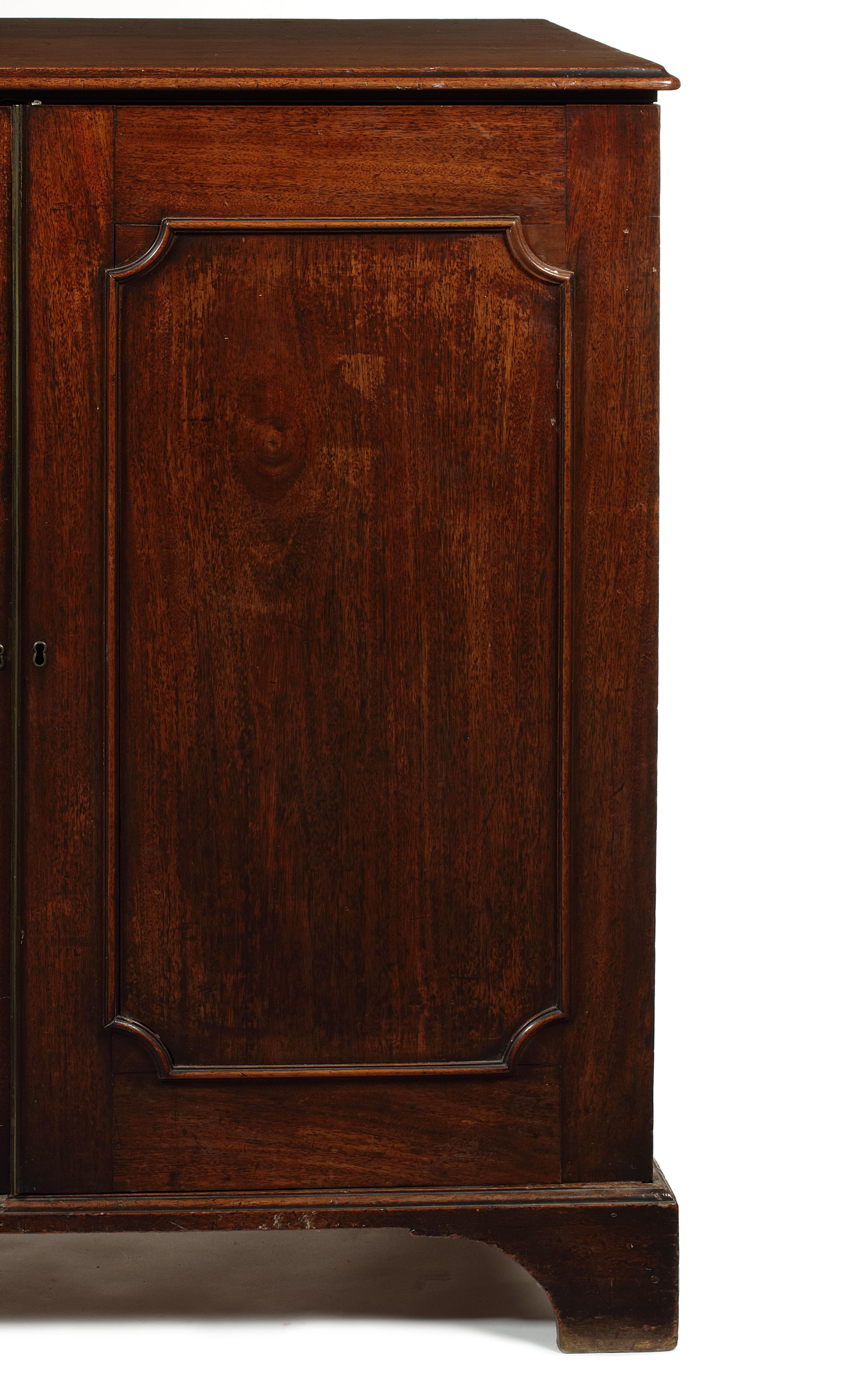 A George III mahogany low press cupboard
The rectangular moulded top above a pair of panelled doors enclosing four slides above two short and one long drawer, on bracket feet, 119 cm wide, 63 cm deep, 118 cm high.
