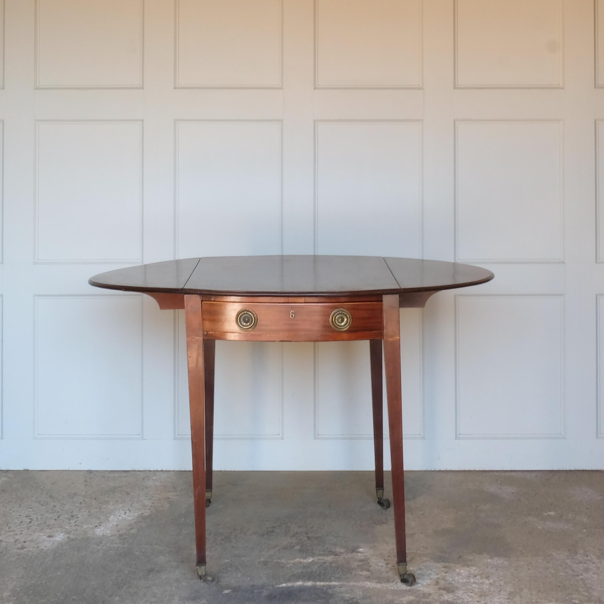 A charming George III mahogany Pembroke table, an oval top with delicate boxwood stringing at the edges over a single drawer, with working key, a dummy drawer matching on the other side, over square tapering legs on brass casters, some minor