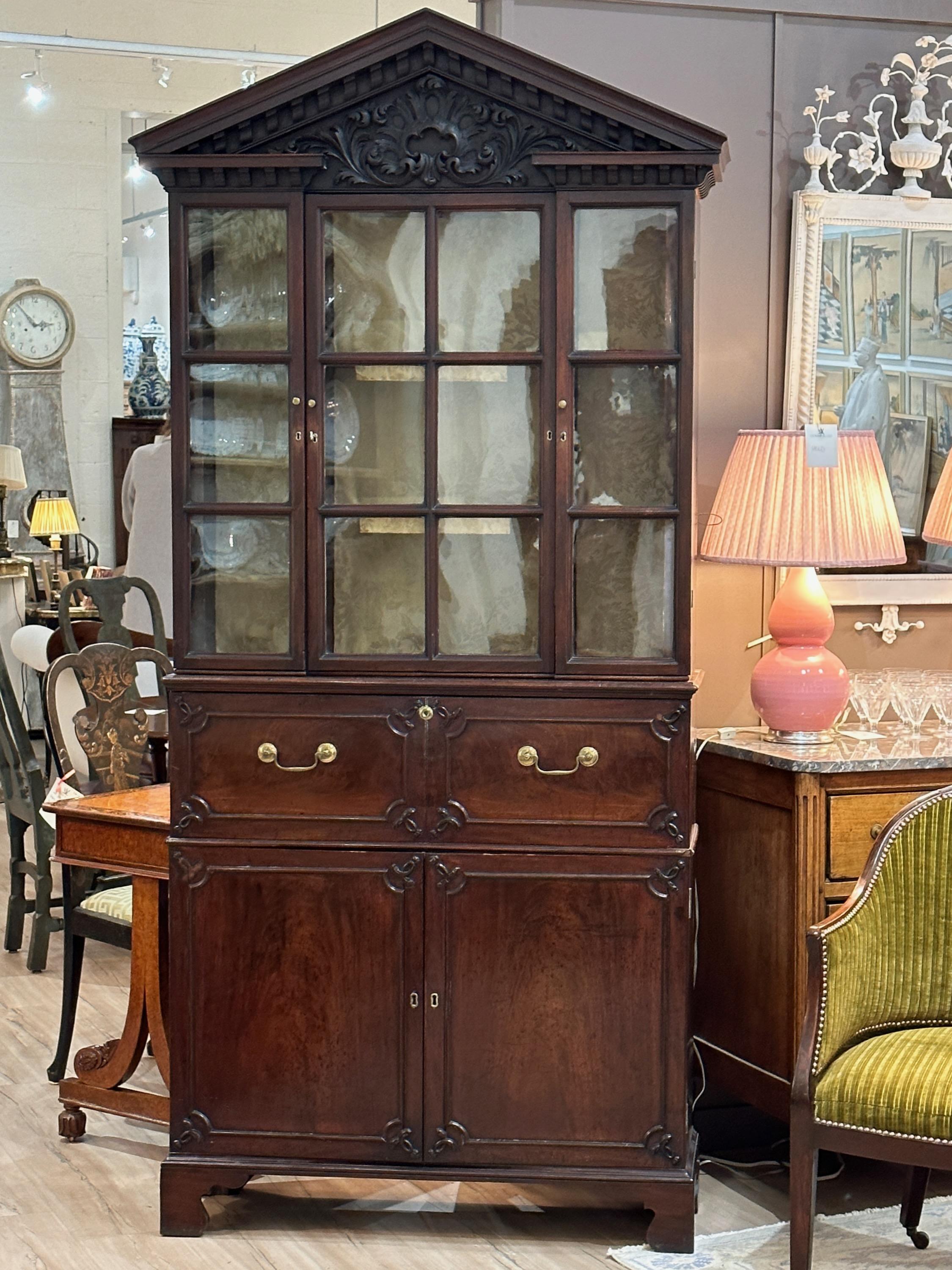 An unusually small and elegant late 18th century carved mahogany secretary bookcase. The intricately carved pediment sits atop a bookcase with three glass fronted doors over a drop front secretary desk with drawers and pigeon holes above two lower