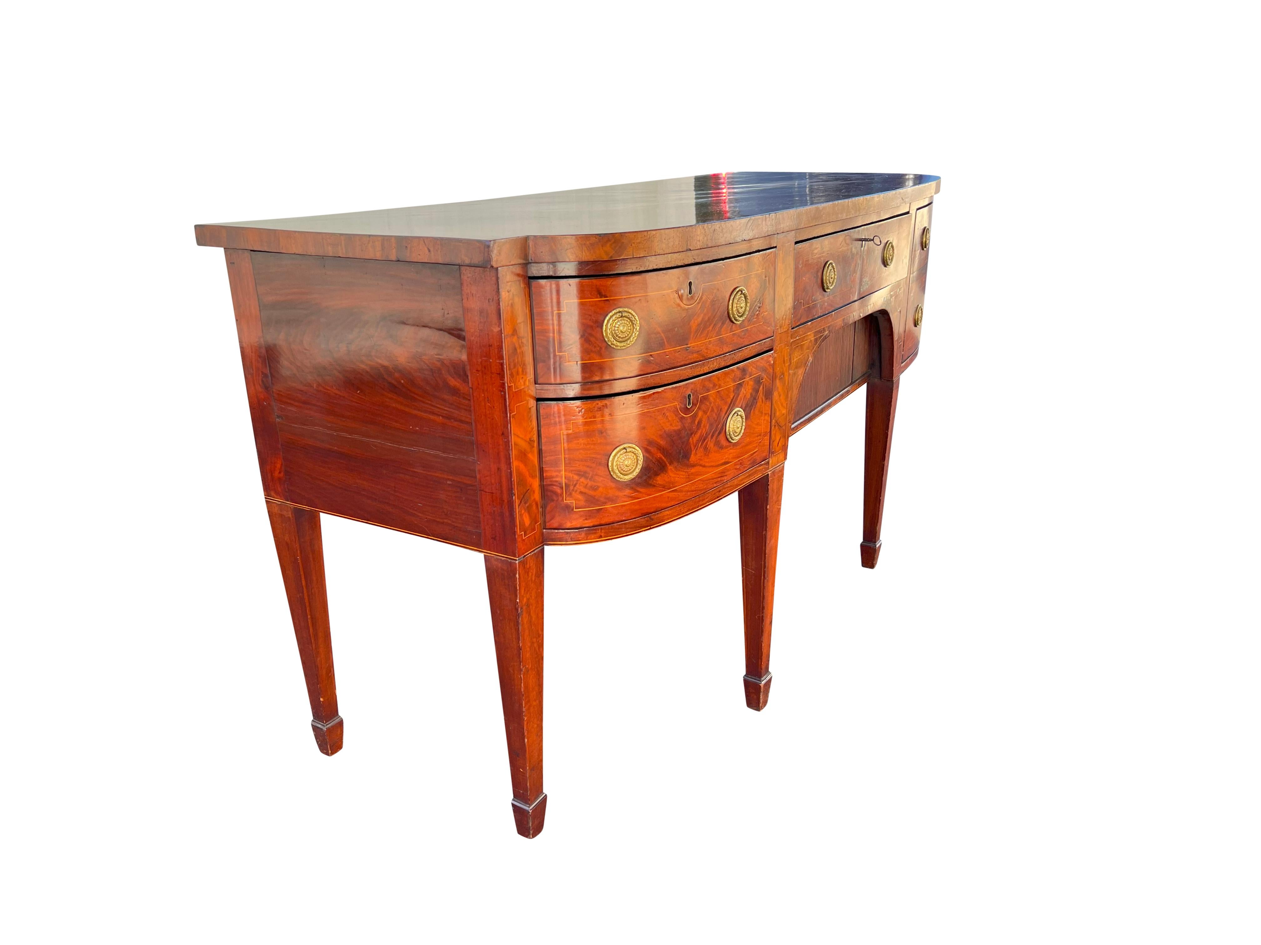 With a rectangular top with rounded ends over a central drawer flanked by a deep bottle drawer and two drawers with tambour door well below. Raised on square tapered legs and spade feet.