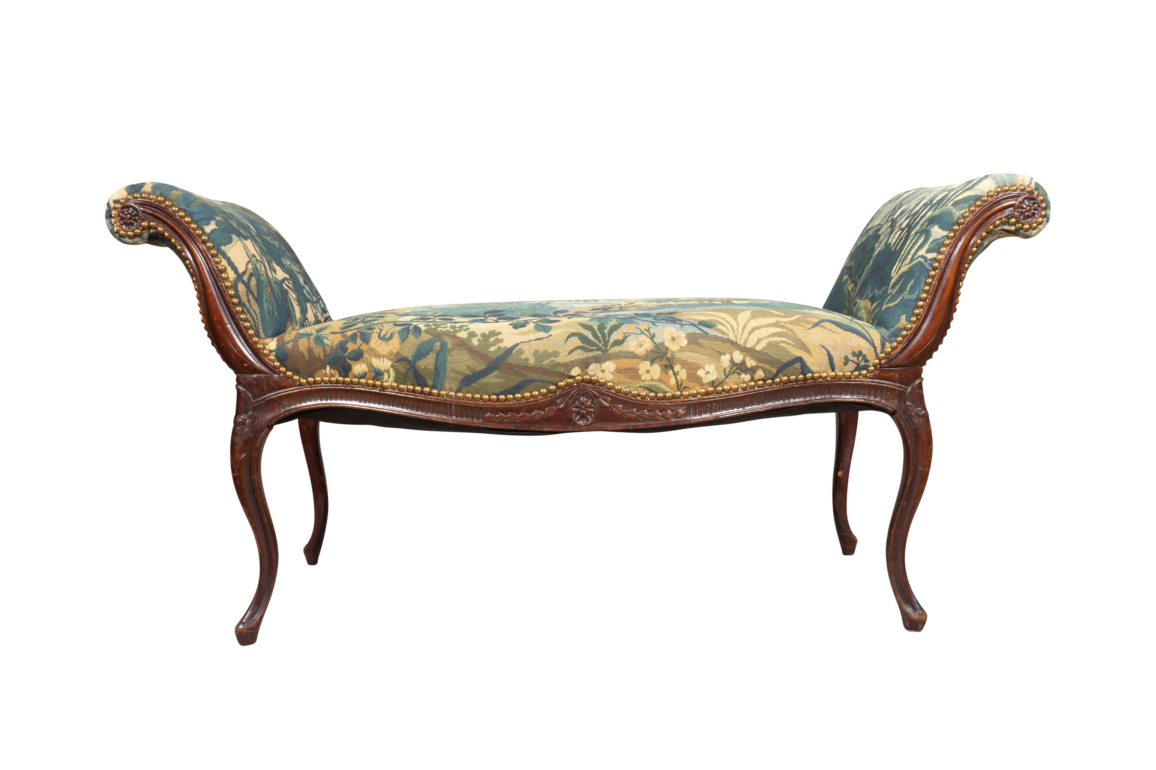In the French taste. With bowed seat upholstered in verdure upholstery with tacks and scrolled ends. The frame carved with central paterae and trailing bell flowers. Paterae at the arm terminals Raised on molded cabriole legs.
