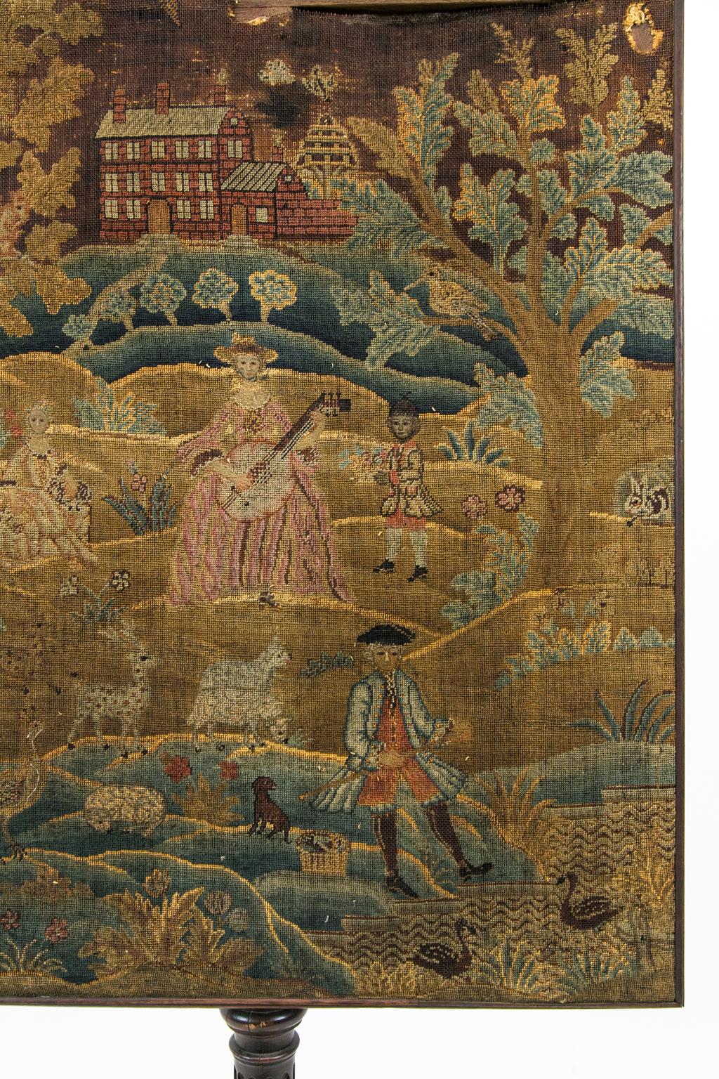 George III needlework fire screen depicts an English manor house with dogs, deer, and peacocks on a landscape. A woman is playing the mandolin while children are playing. The faces of the figures are stitched in raised petit point. The stem is