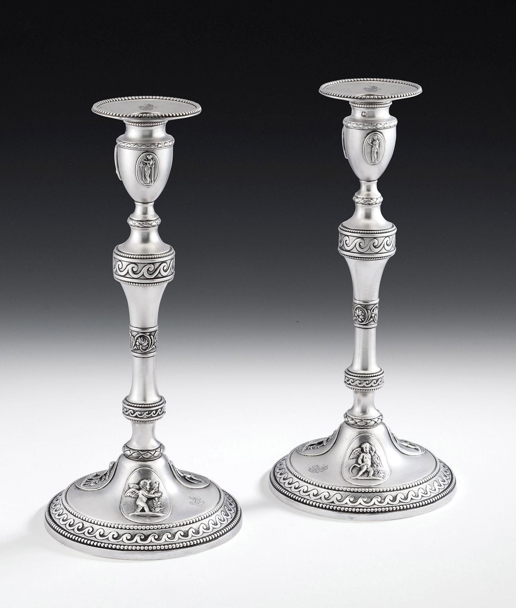 These exceptionally rare cast candlesticks have a circular base, with a double beaded edge, enclosing a band of wave motifs on a matted ground. Most unusually the base is decorated with raised cast classical medallions depicting Cupid with the