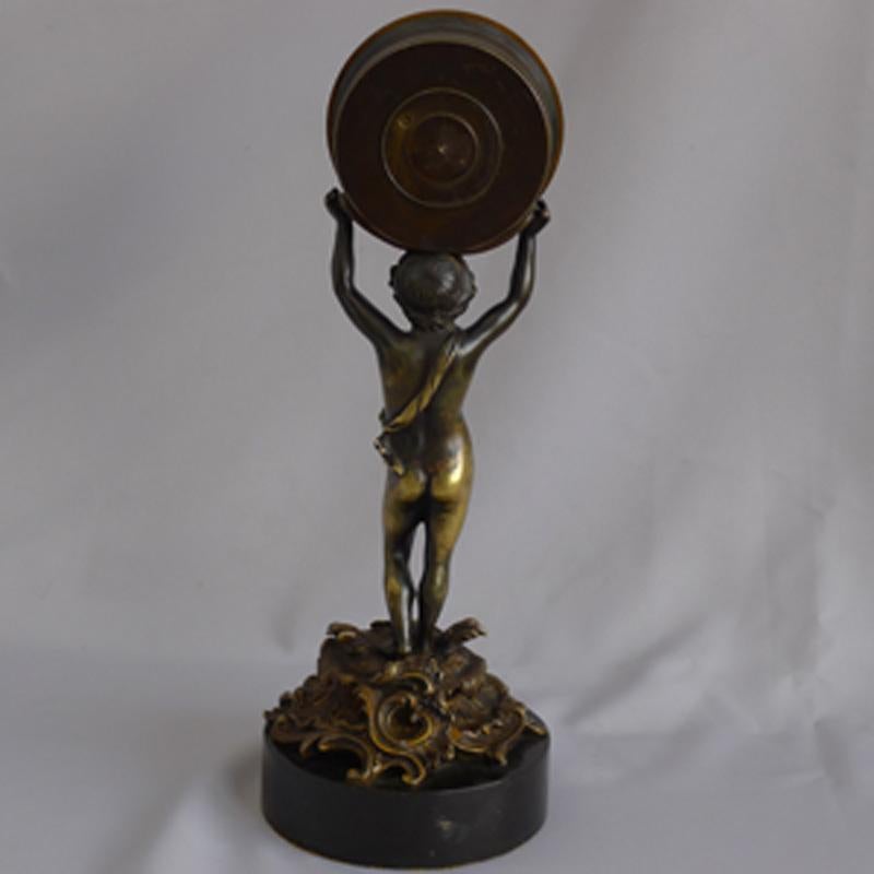 A superb quality and rare George III patinated bronze and ormolu figural mantel clock by W. Glover of chronometer quality. Set on a circular black marble base, the putti stands on a scolling, rocco style ormolu raised base. The putti has a
