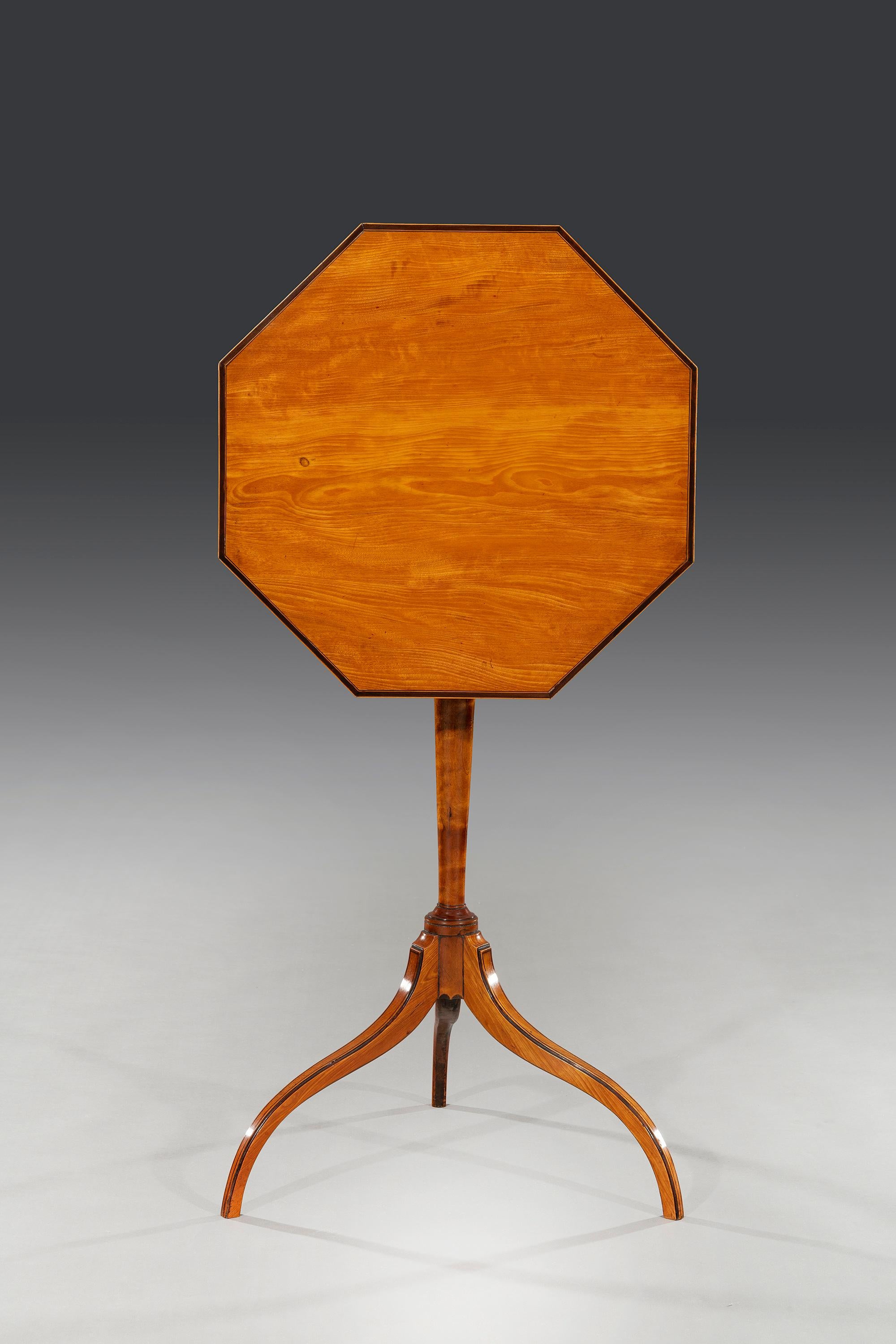 The octagonal tilt-top table has purple-heart cross banding and is veneered in figured West Indian satinwood and made through-out in solid satinwood. The table stands on a tripod base with elegant turned pedestal column and down-swept inlaid tapered