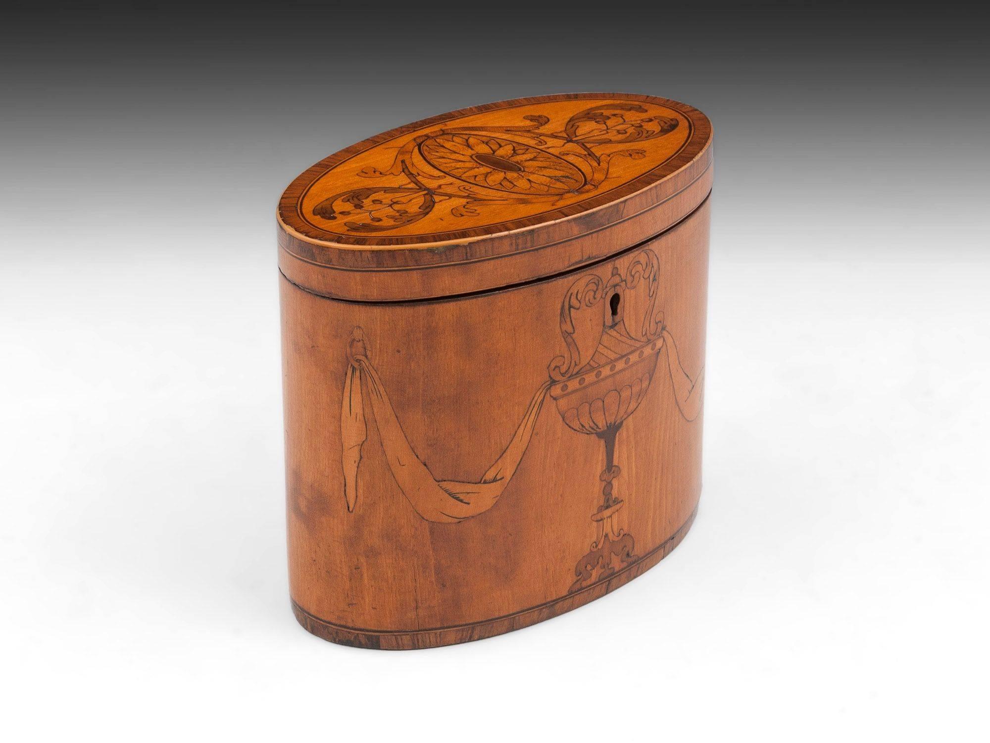 The tea caddy has kingwood crossbanding and boxwood edging and is inlaid with Adam style urn and husk swags. The top has symmetrical entwined leaves surrounding an inlaid oval paterae.