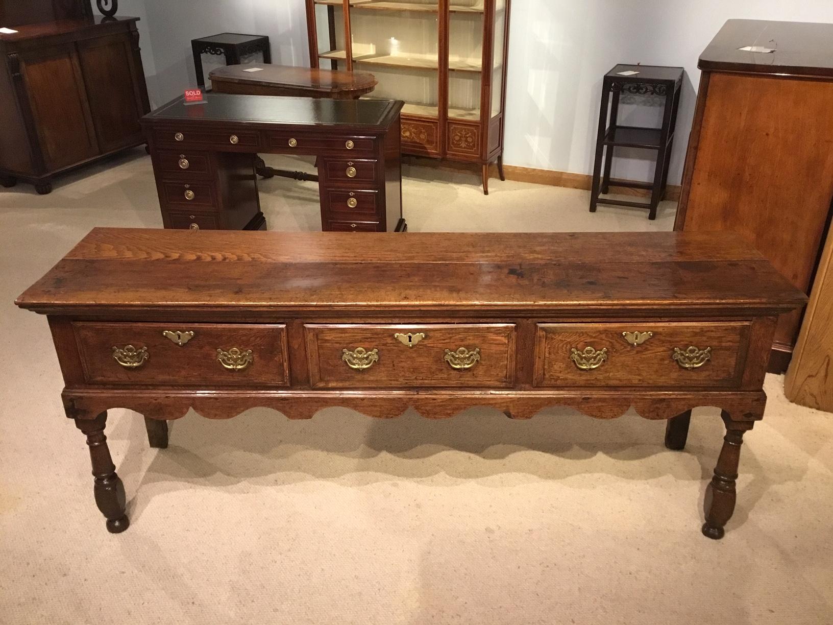 A good George III period antique oak dresser base. The rectangular two plank top above three rectangular oak drawers with brass swan neck handles and escutcheons. Having a wavy shaped apron and raised on turned front supports and stile rear supports