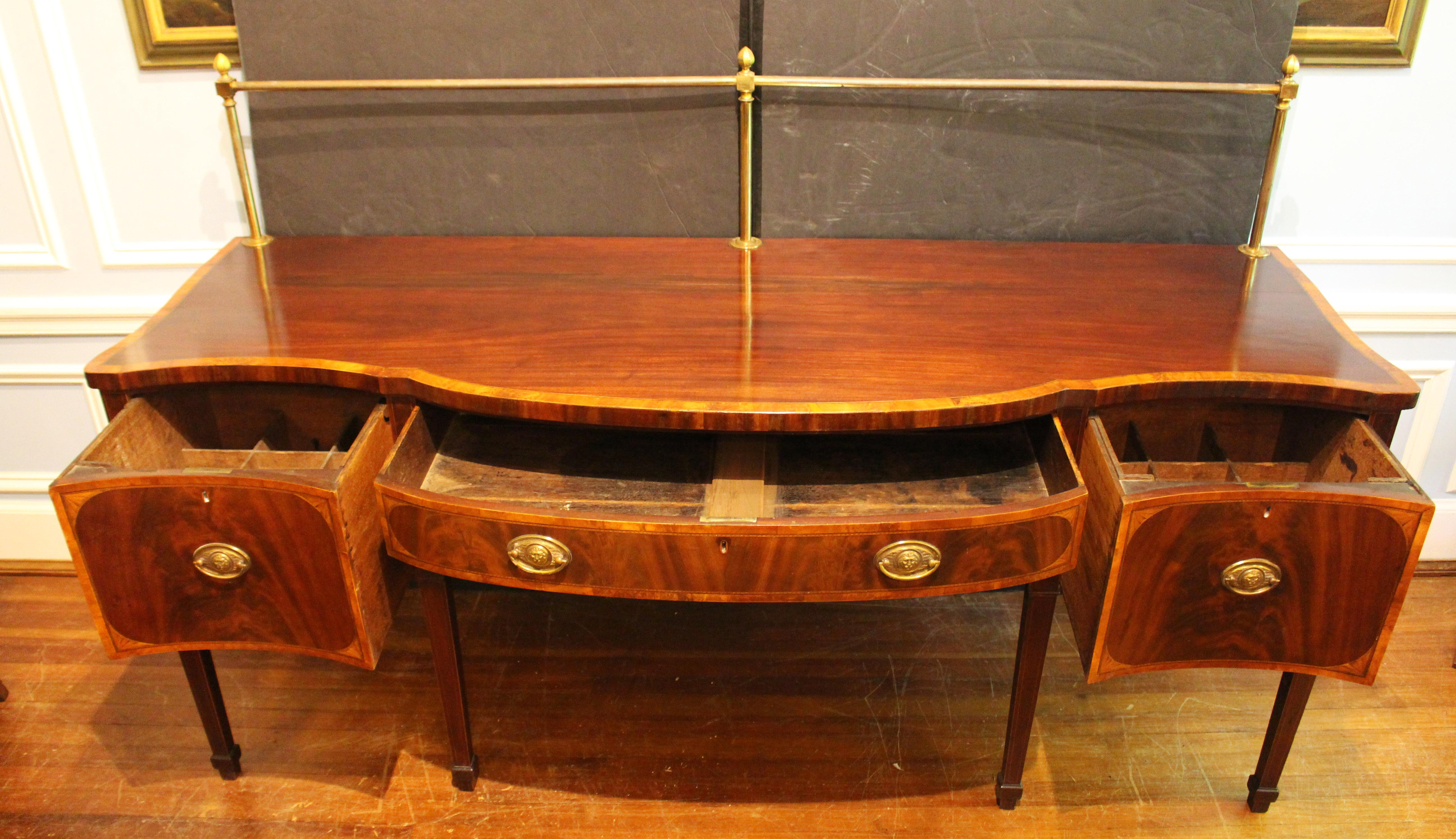 George III period serpentine front sideboard with brass rail, English. Circa 1780. Oval lion mask brass pulls. Central long drawer over stunning sun inlaid spandrels; flanked by two deep cellarette drawers. The top and each drawer front with
