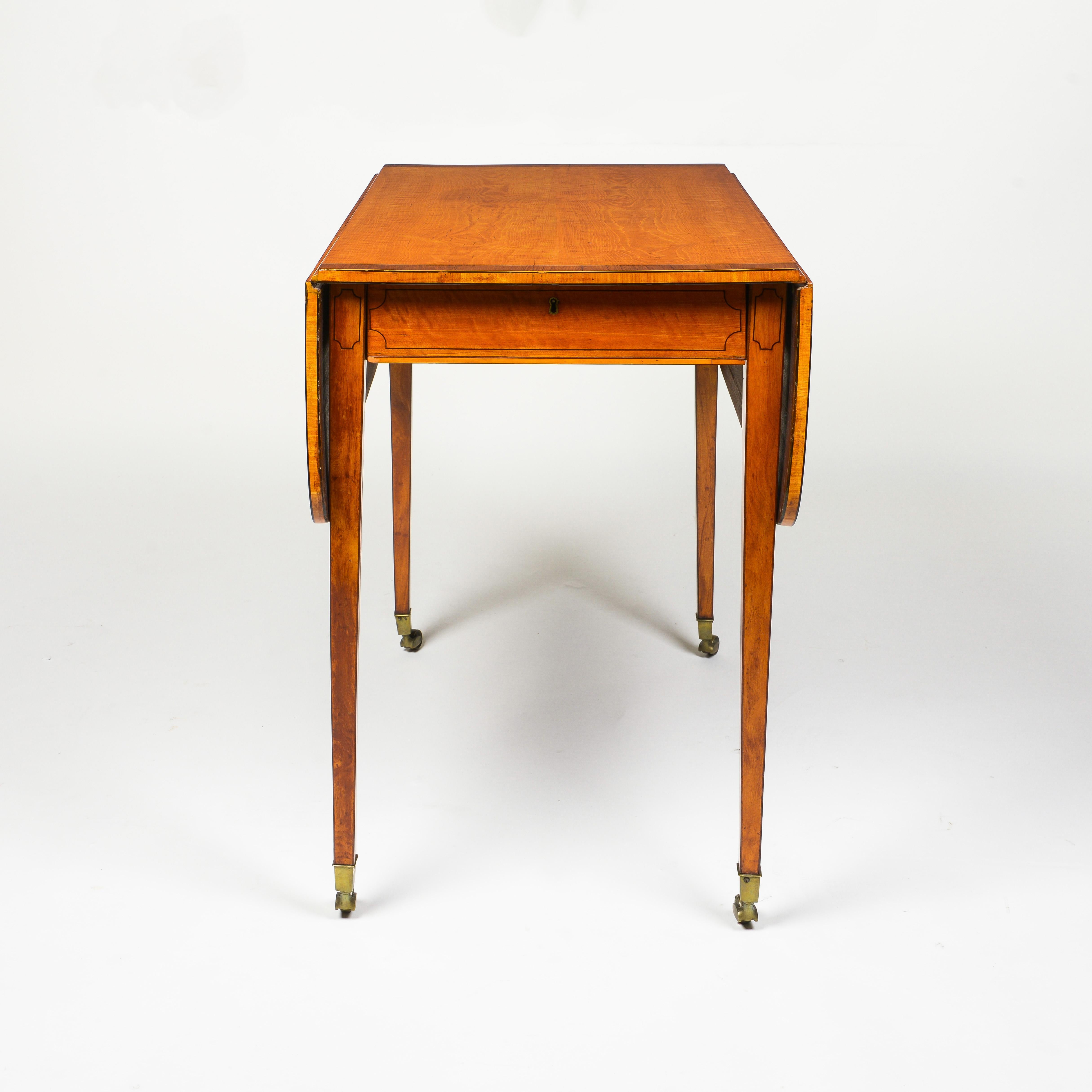 With ebonized stringing throughout; the rectangular mahogany banded top with drop leaves over a frieze drawer, raised on square tapering legs ending in brass caps and casters.